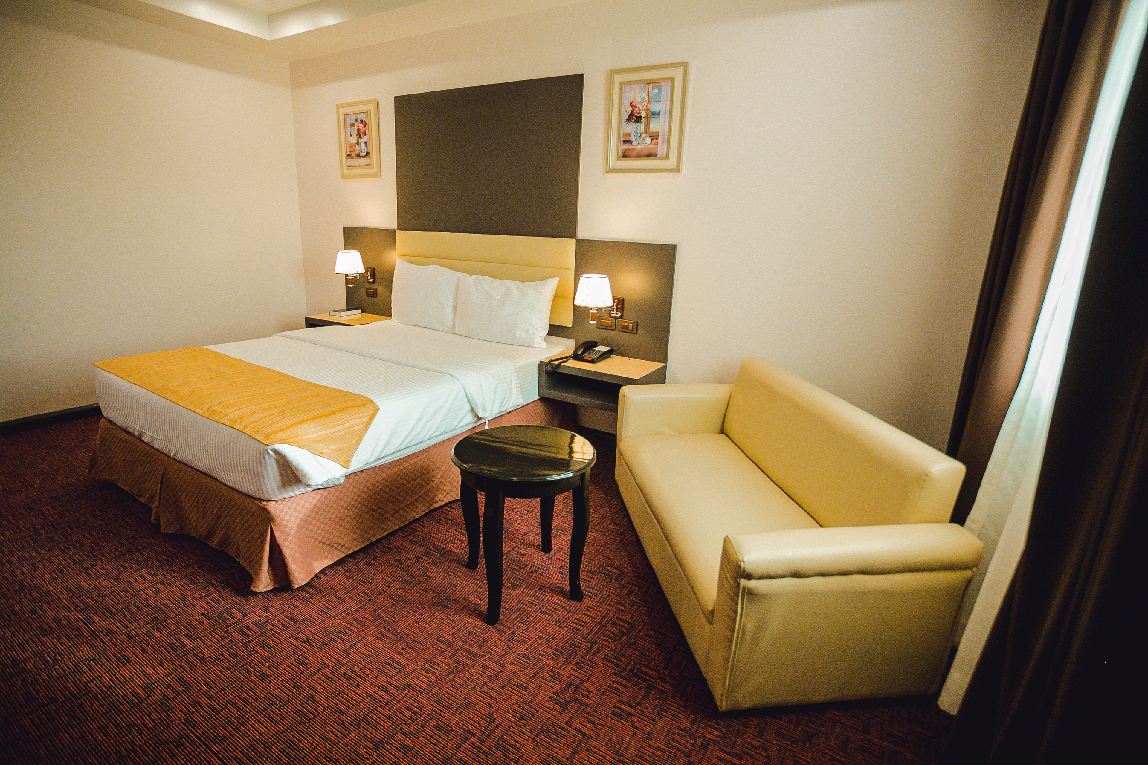 A hotel room with a bed, a nightstand, a lamp, a sofa, and a small table.