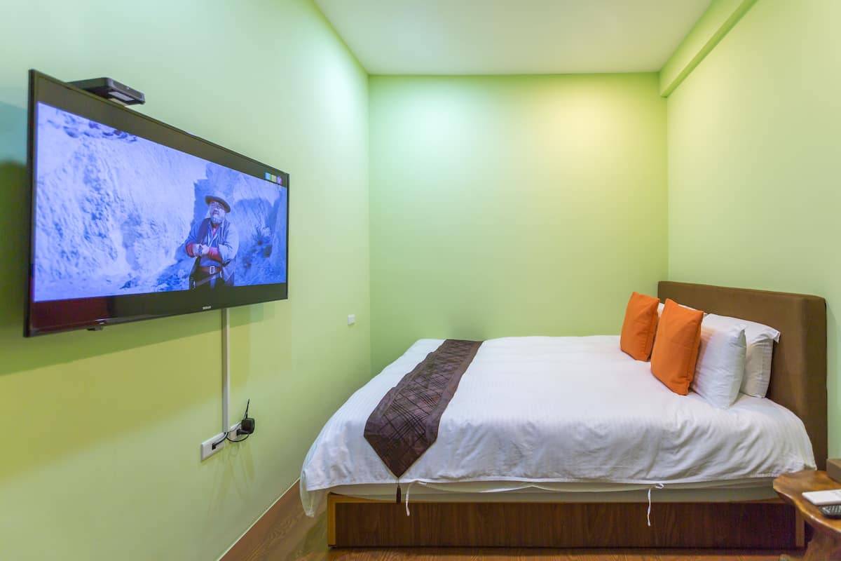 Una habitación de hotel con cama, televisor montado en la pared y mesita de noche de madera.