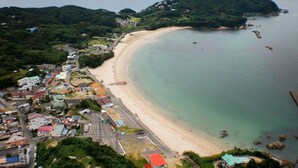 Vue aérienne d’une zone côtière avec une plage, des bâtiments et une chaîne de montagnes en arrière-plan.