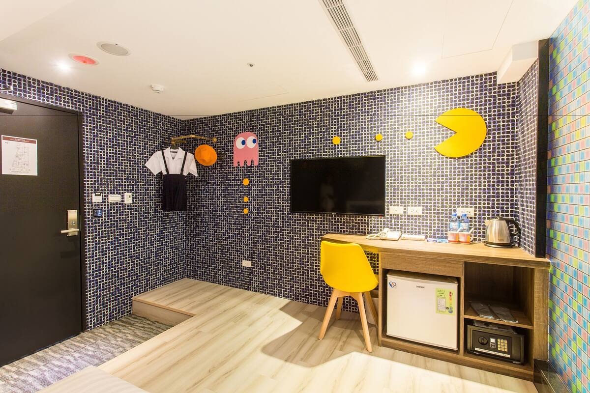 A room with a tiled wall, a flat-screen TV, a wooden desk, a yellow chair, and a small refrigerator.