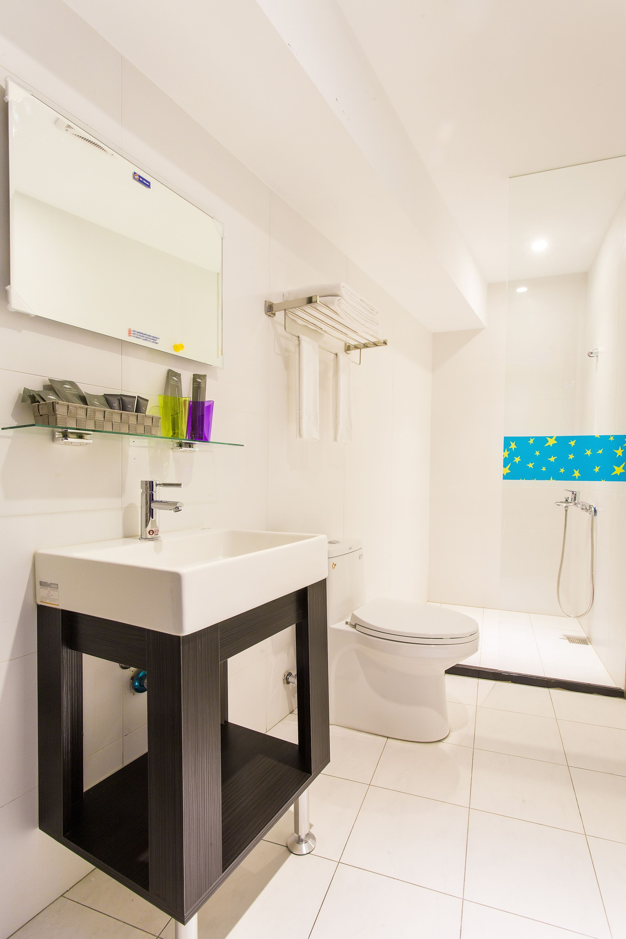 A modern bathroom with a white sink, mirror, and toilet. There is a glass shelf above the sink holding toiletries.