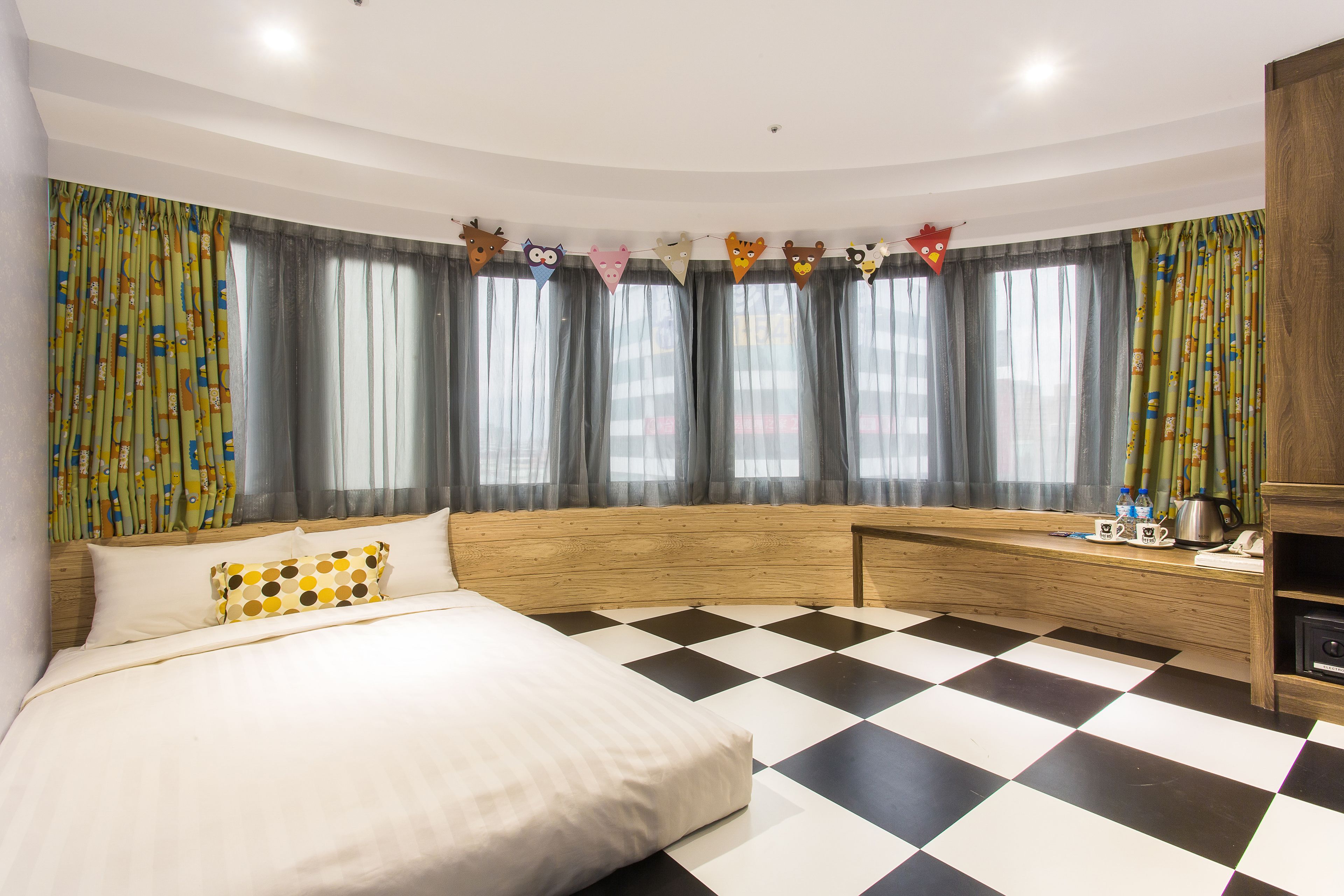 A hotel room with a checkered floor, a bed with white and yellow pillows, a wooden headboard, and a desk with a kettle and cups.