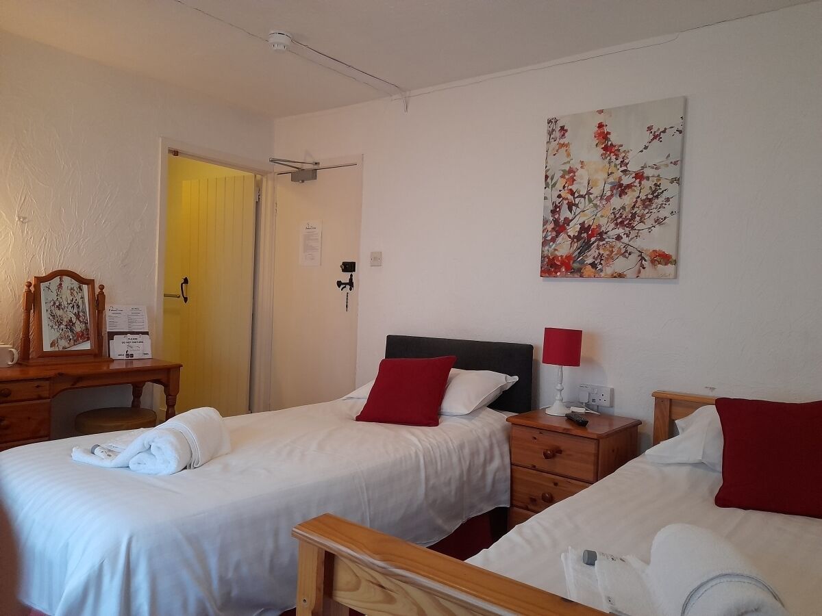 A hotel room with two single beds, a wooden desk, a chair, a nightstand with a lamp, and a painting on the wall.