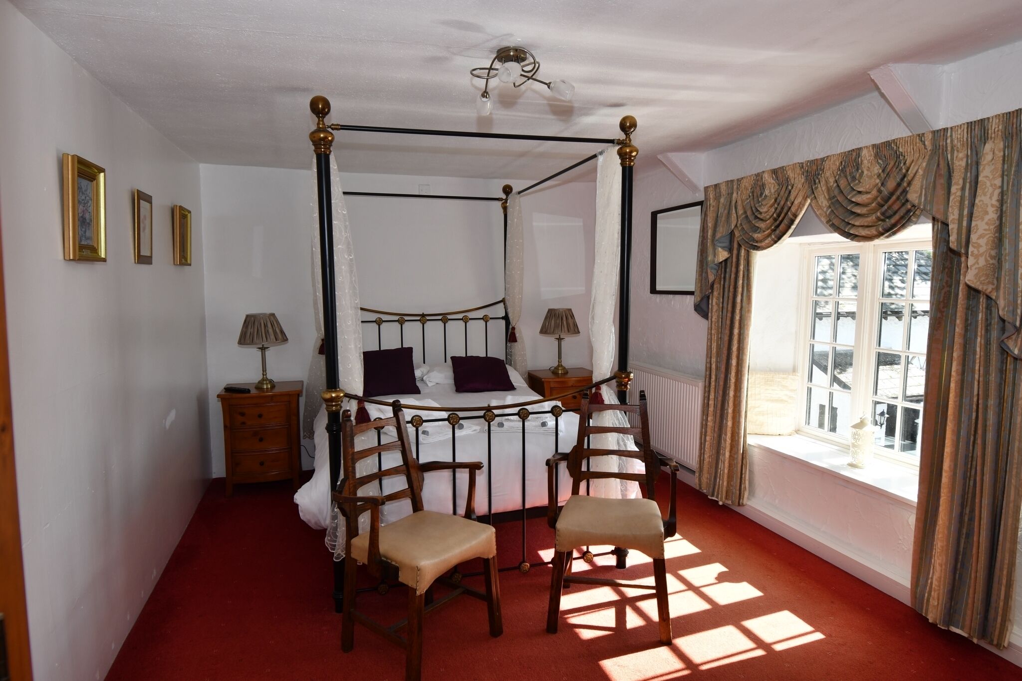 Una cama con dosel y cortinas, sillas de madera, una mesita de noche y una ventana con cortinas.