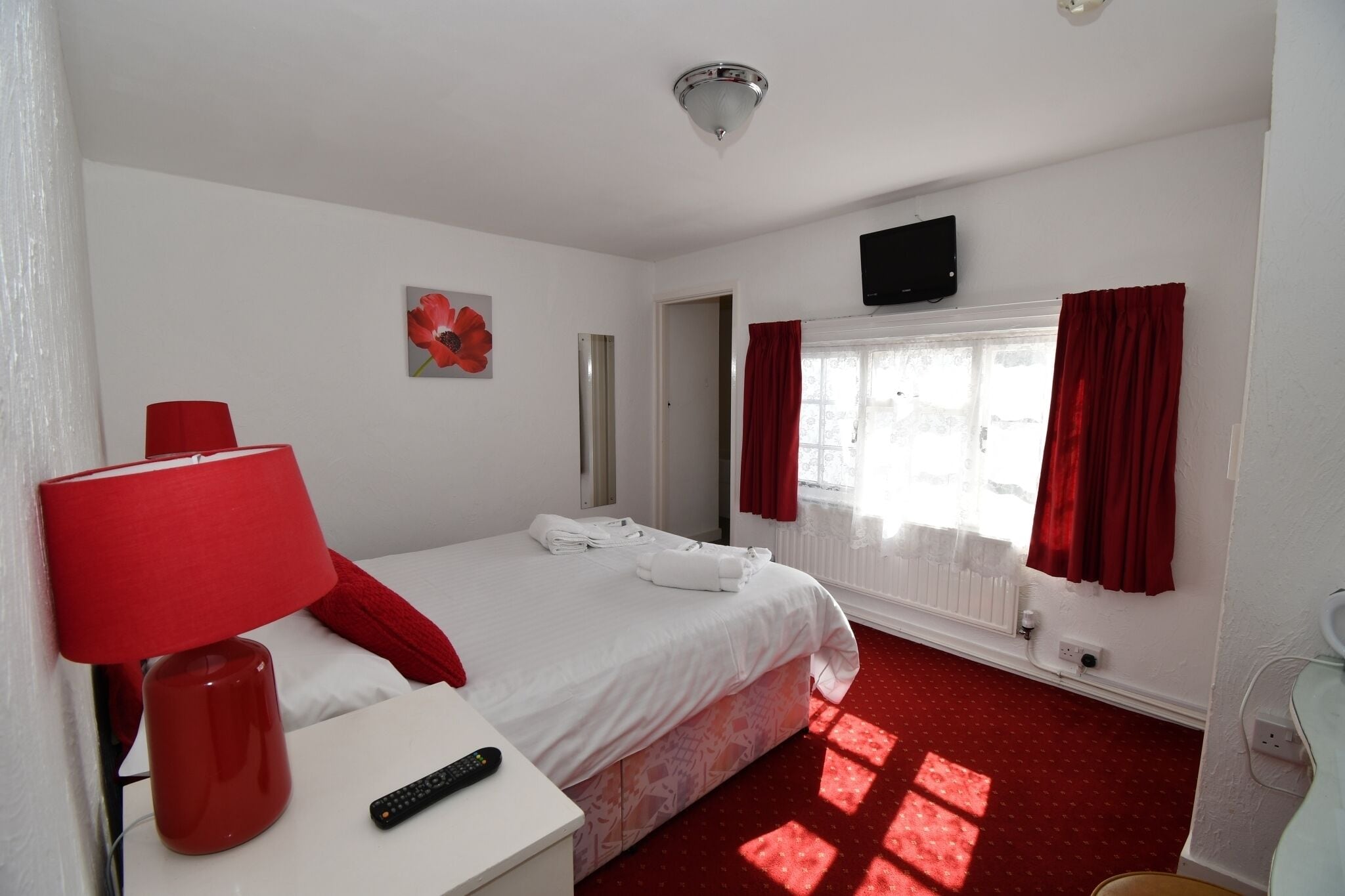 Una habitación de hotel con una cama, una lámpara roja, un televisor y una ventana con cortinas rojas.