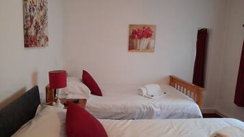 A hotel room with two single beds, a wooden headboard, a red lamp, a white pillow, and a painting of flowers on the wall.