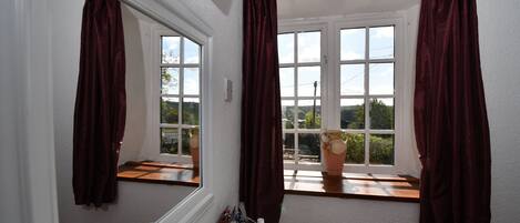 A bathroom with a large mirror, two windows with curtains, a sink with toiletries, and a view of the outdoors.