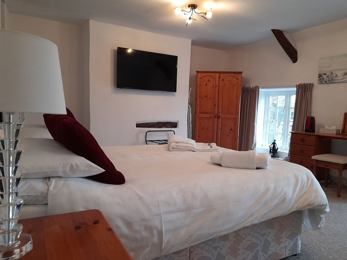 Ein ordentlich eingerichtetes Schlafzimmer mit einem großen Bett, einem flachbildfernseher an der Wand, einem Holzschrank und einem Schreibtisch mit Stuhl.
