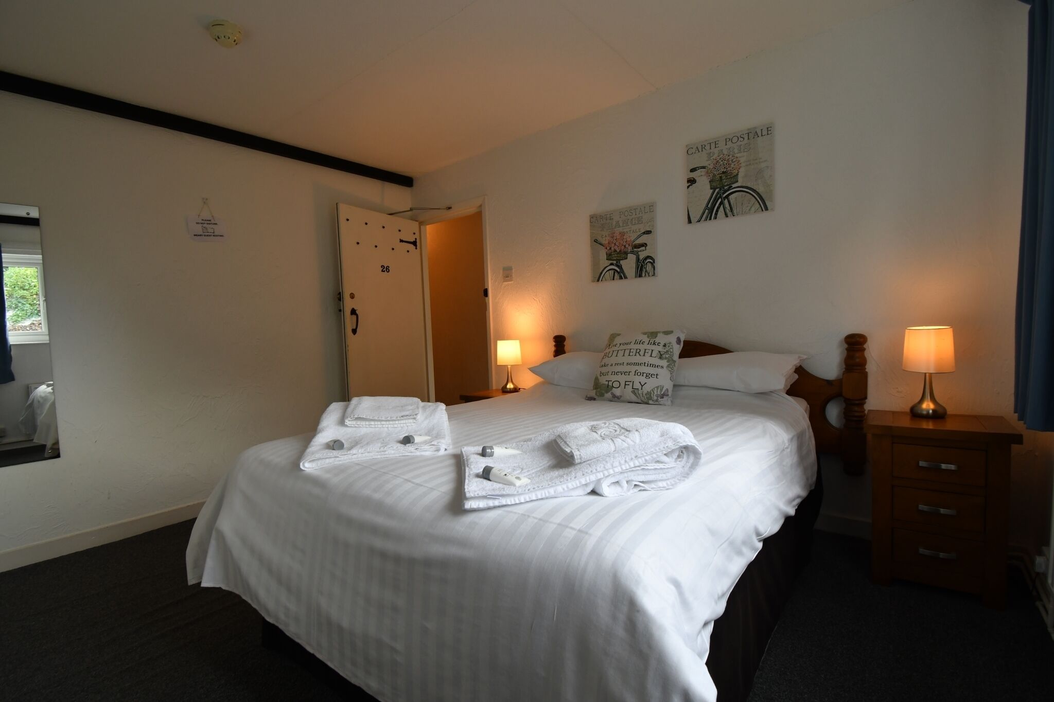 A neatly made bed with white linens and towels, a wooden nightstand with a lamp, a door with a sign, and a wall with a bicycle illustration and a sign reading "CARTE POSTALE".
