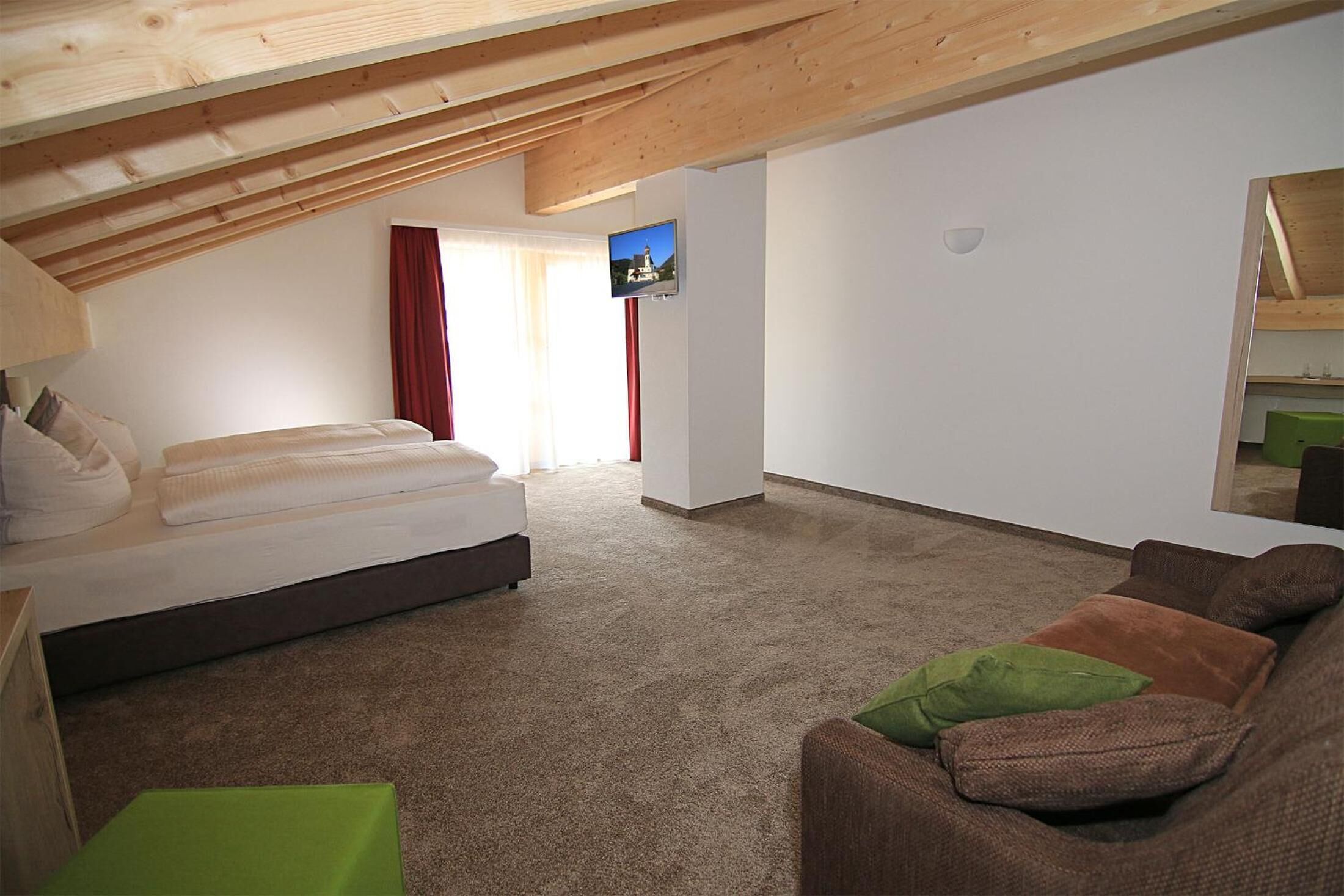 A bedroom with a bed, a green ottoman, a brown sofa, a TV on the wall, and a wooden ceiling.