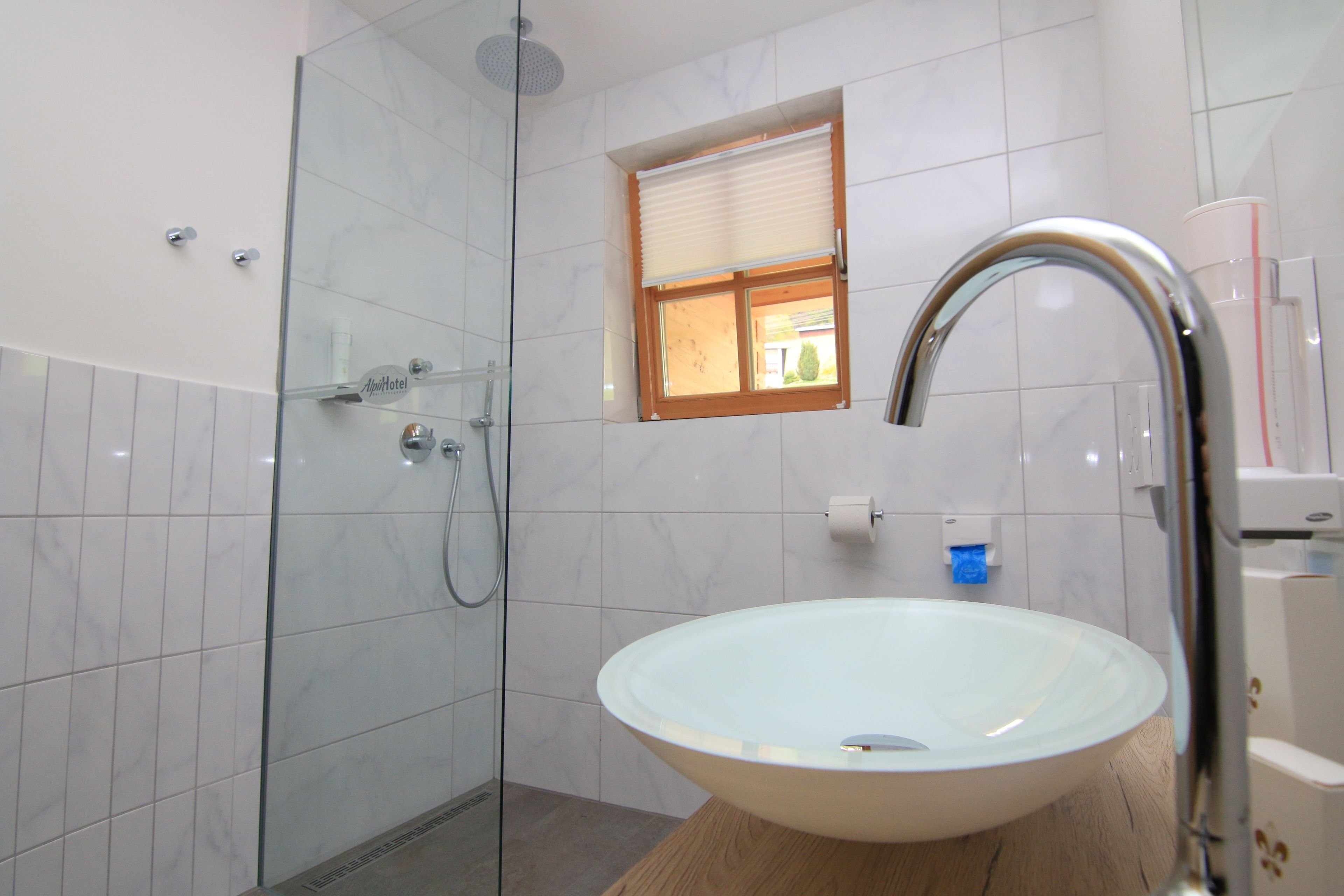 Une salle de bain moderne avec une douche à parois de verre, un lavabo blanc doté d’un robinet chromé et une petite fenêtre avec des stores.