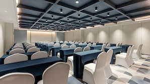 Une salle de conférence avec des rangées de tables et de chaises, un design de plafond moderne et une palette de couleurs neutres.