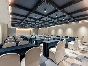 Un salón de conferencias con filas de mesas y sillas, un diseño moderno de techo y una paleta de colores neutros.