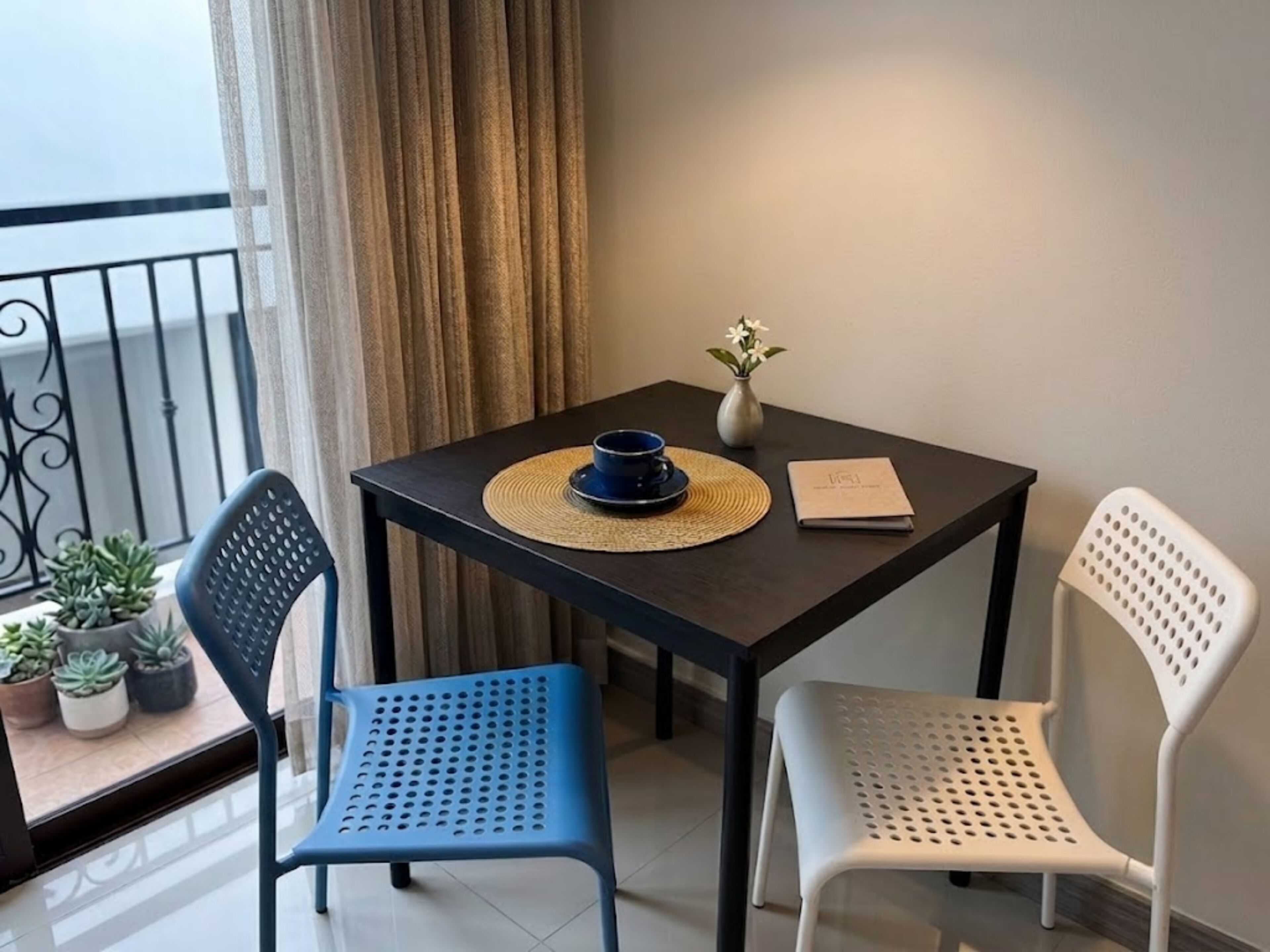 Une petite table sur laquelle se trouvent un bol bleu, un vase blanc avec des fleurs et un livre. On y voit deux chaises, une bleue et une blanche, avec un balcon et des plantes en pot en arrière-plan.