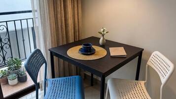 Une petite table sur laquelle se trouvent un bol bleu, un vase blanc avec des fleurs et un livre. On y voit deux chaises, une bleue et une blanche, avec un balcon et des plantes en pot en arrière-plan.