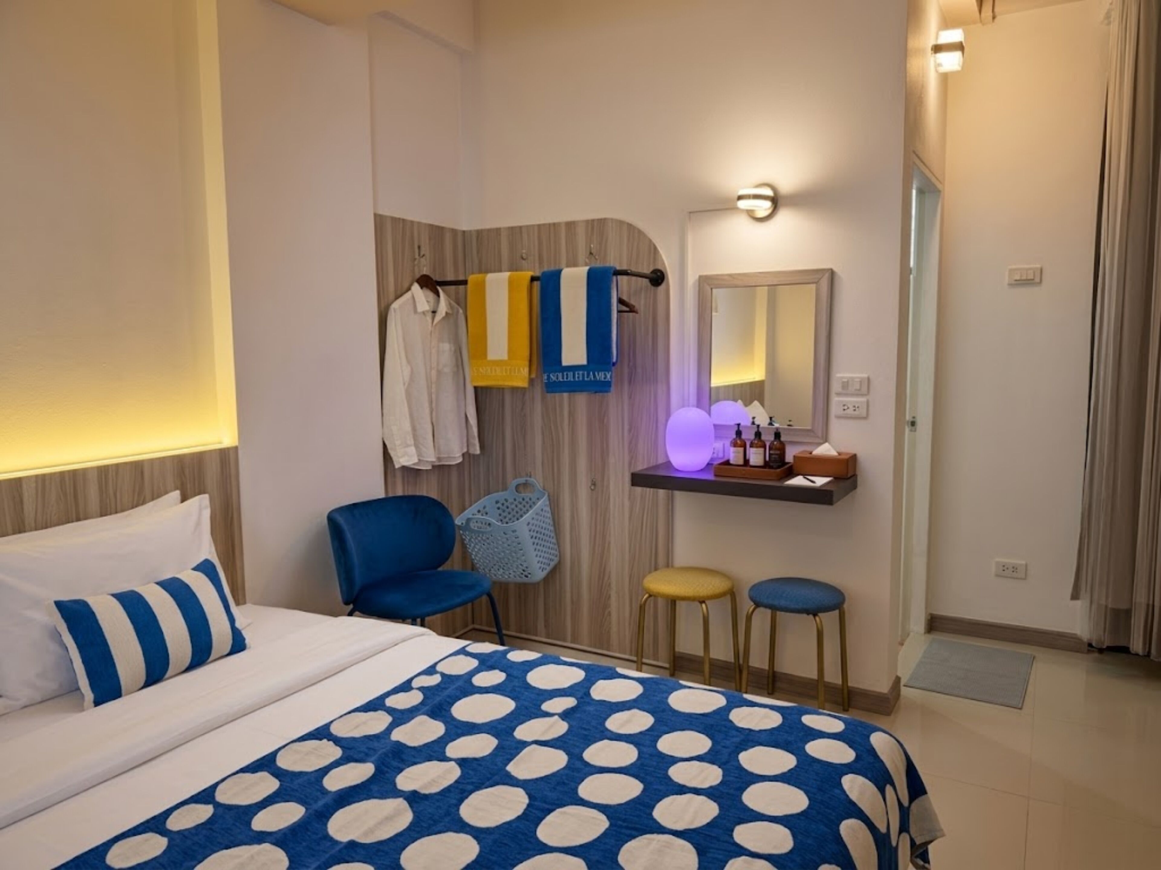 Una habitación de hotel con una cama, una silla, un taburete, una tocador con espejo y un perchero para toallas.