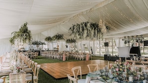 Un espace événementiel intérieur aménagé avec des tables dressées pour les repas, une scène et des décorations florales suspendues.