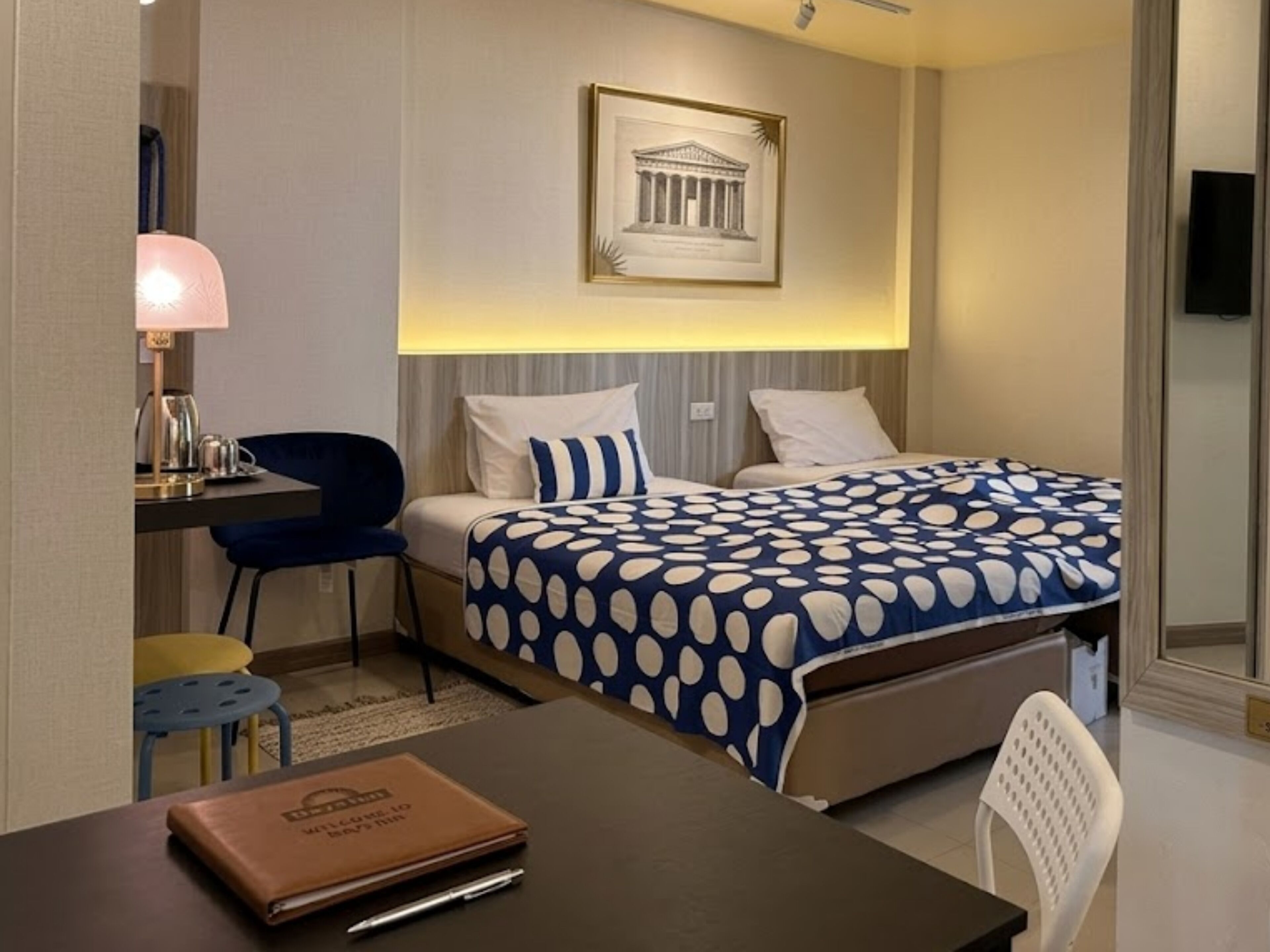 A hotel room with a bed, a desk with a lamp, a chair, a framed picture on the wall, and a small table with a book.