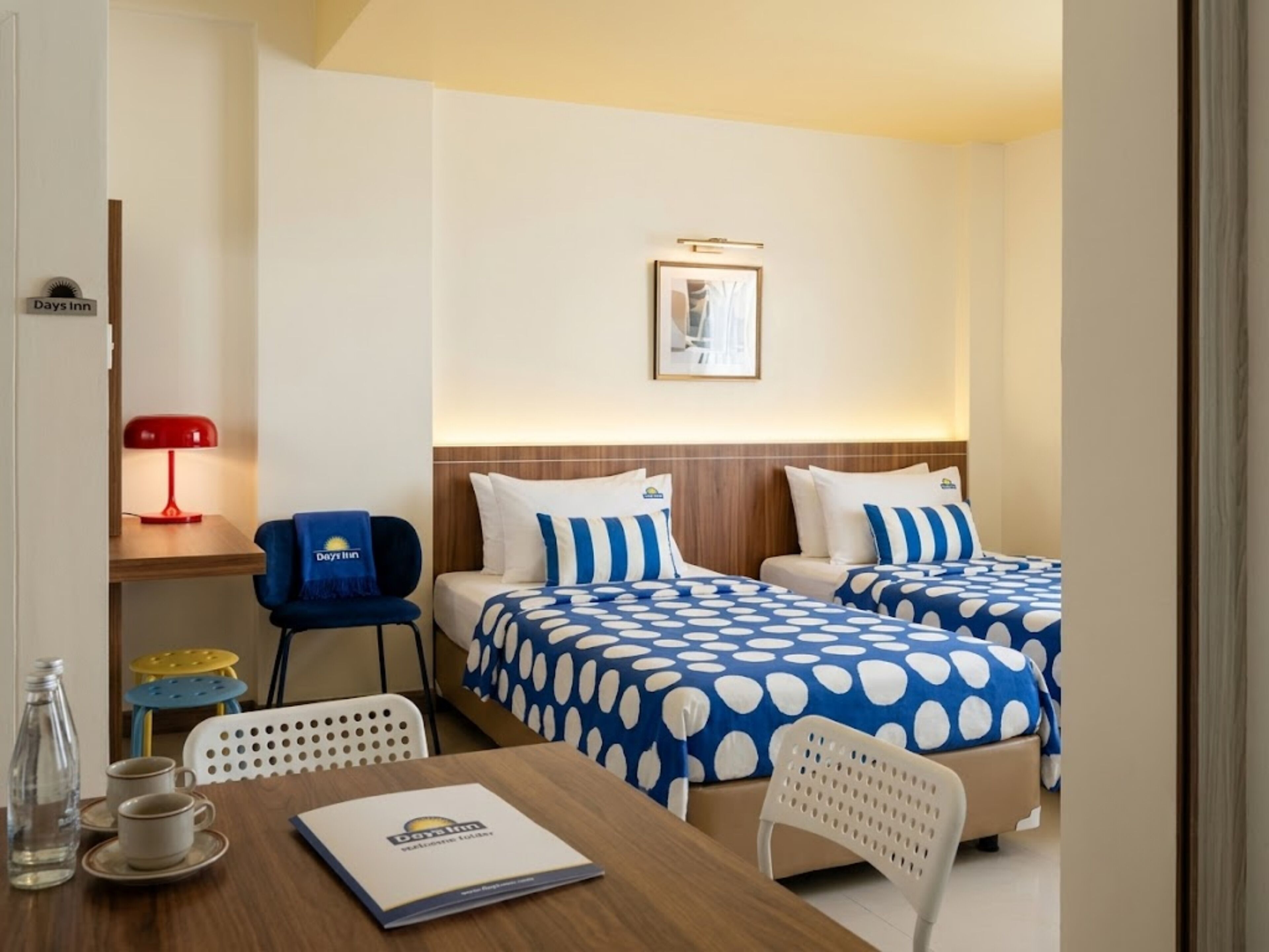 Une chambre d’hôtel avec deux lits, un bureau doté d’une lampe rouge, une chaise, une table sur laquelle se trouve une brochure, et un tableau encadré accroché au mur.