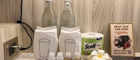 Un baño con encimera de mármol, dos botellas de vidrio, un rollo de papel higiénico, un florero y un cartel que anuncia alquiler de coches.