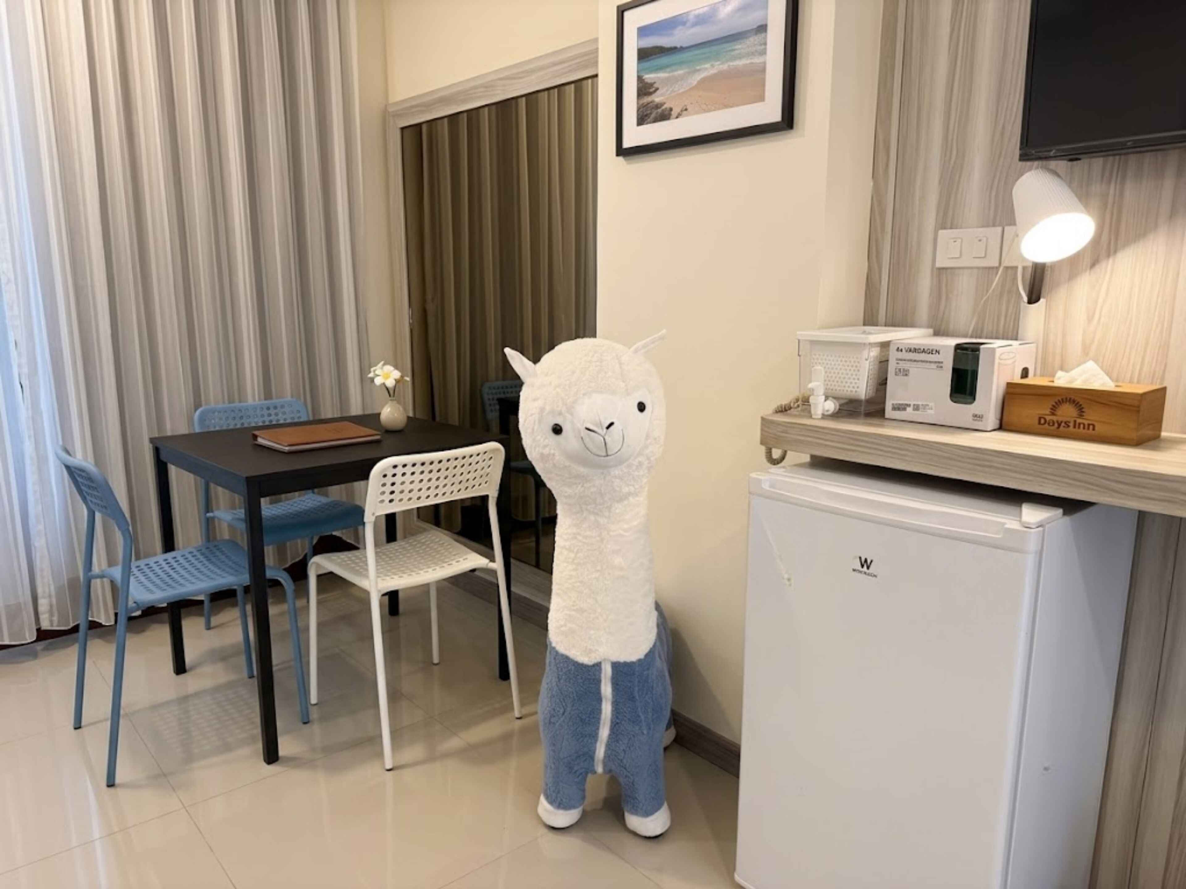 Habitación de hotel con comedor, un refrigerador pequeño, una lámpara y un peluche.