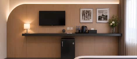 Una habitación de hotel moderna con una televisión de pantalla plana, un refrigerador pequeño, una mesita de noche con lámpara y obras de arte en la pared.