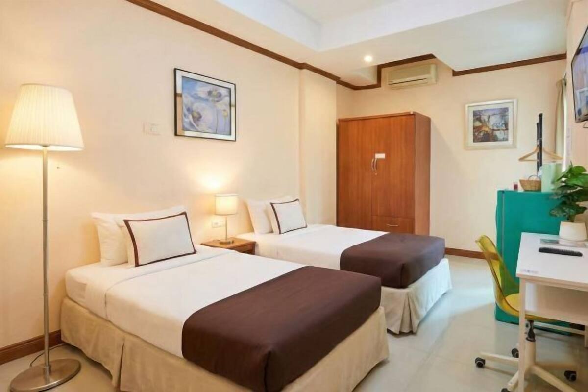 Habitación de hotel con dos camas, un escritorio, una silla, un armario y una lámpara.