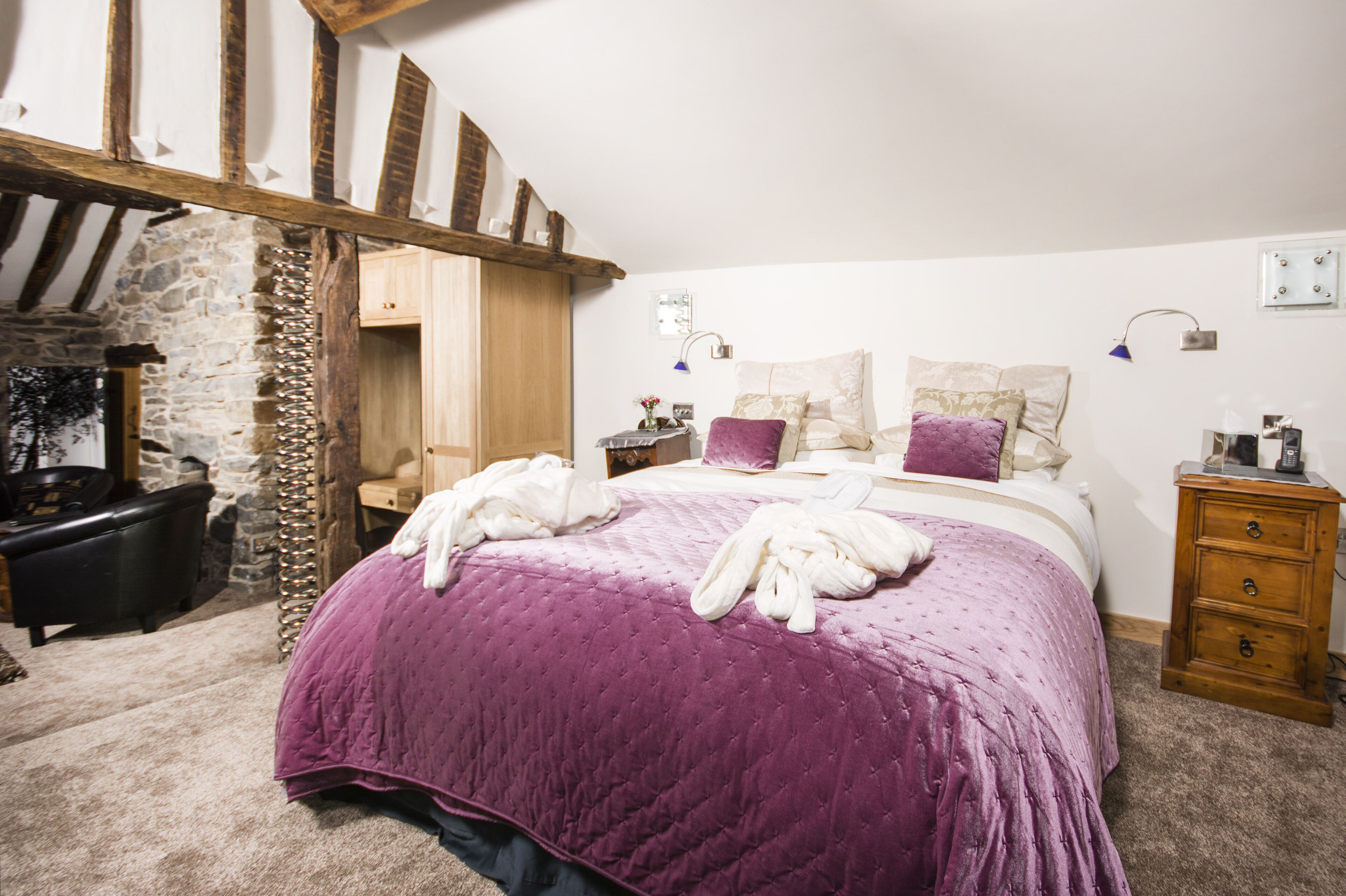 Un dormitorio con una cama grande, pared de piedra, vigas de madera, cómoda de madera y mesita de noche.