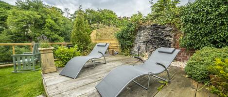 A wooden deck with two reclining chairs and a small table, surrounded by lush greenery and a stone wall.