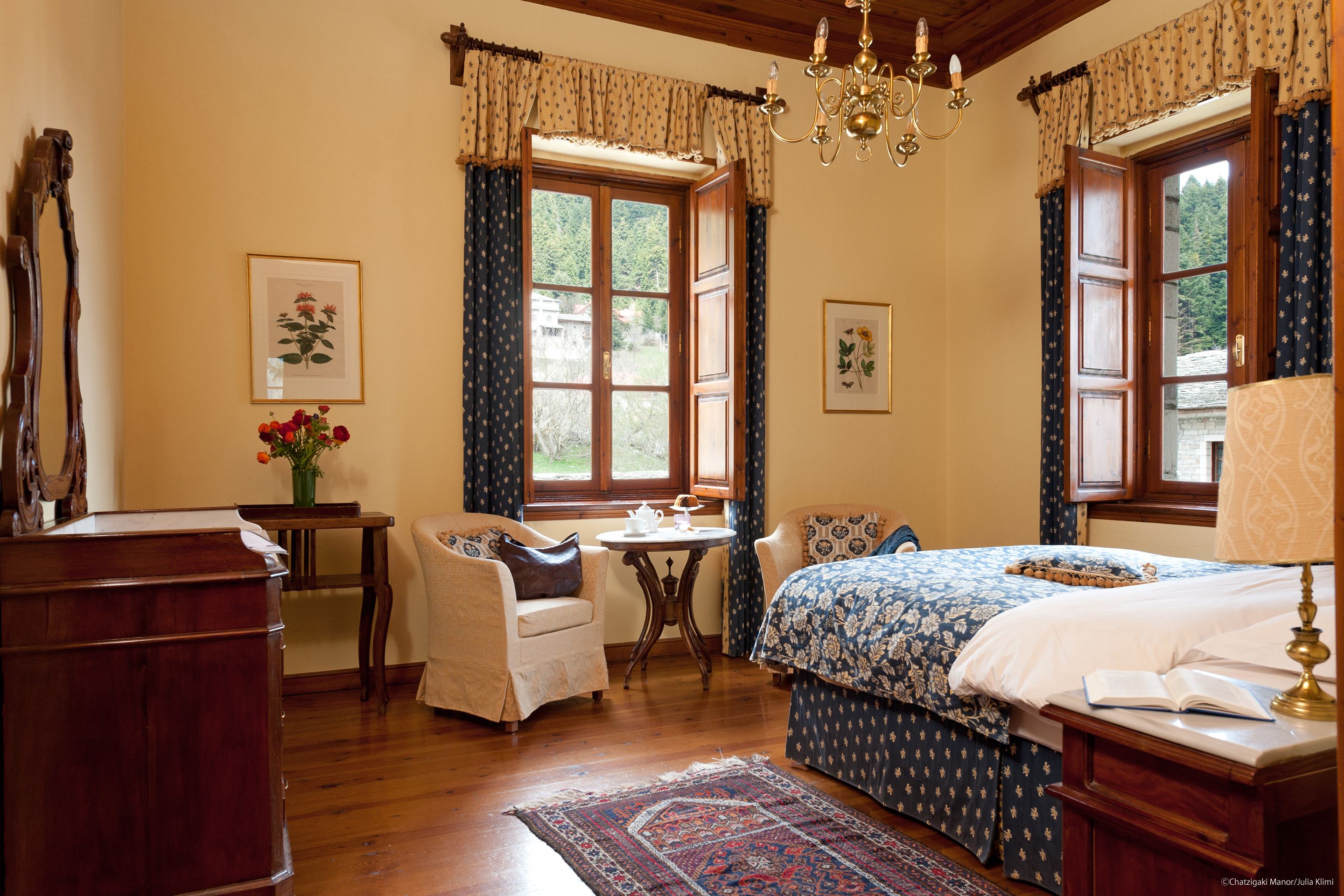 A bedroom with a large bed, a wooden dresser, a small table with a vase of flowers, and a chair.