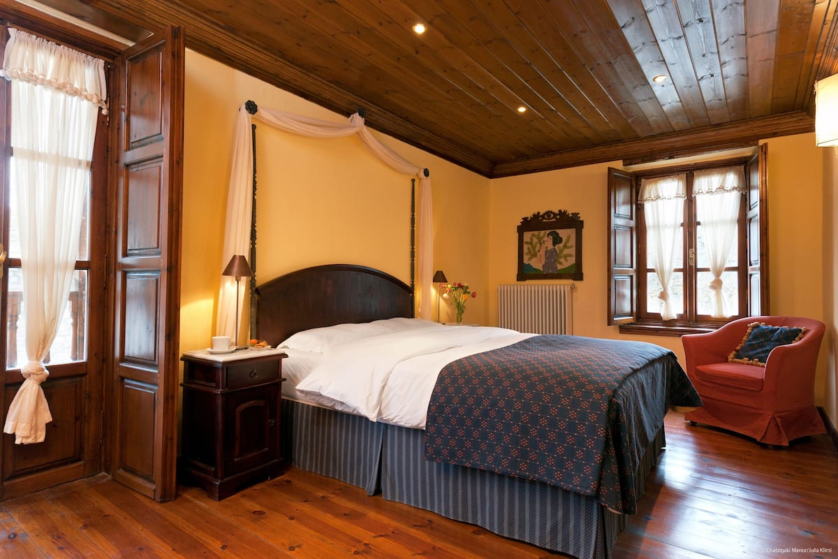 Ein Schlafzimmer mit Holzboden, einem Himmelbett, einem Nachttisch mit Lampe, einem Sessel und einem Fenster mit Vorhängen.