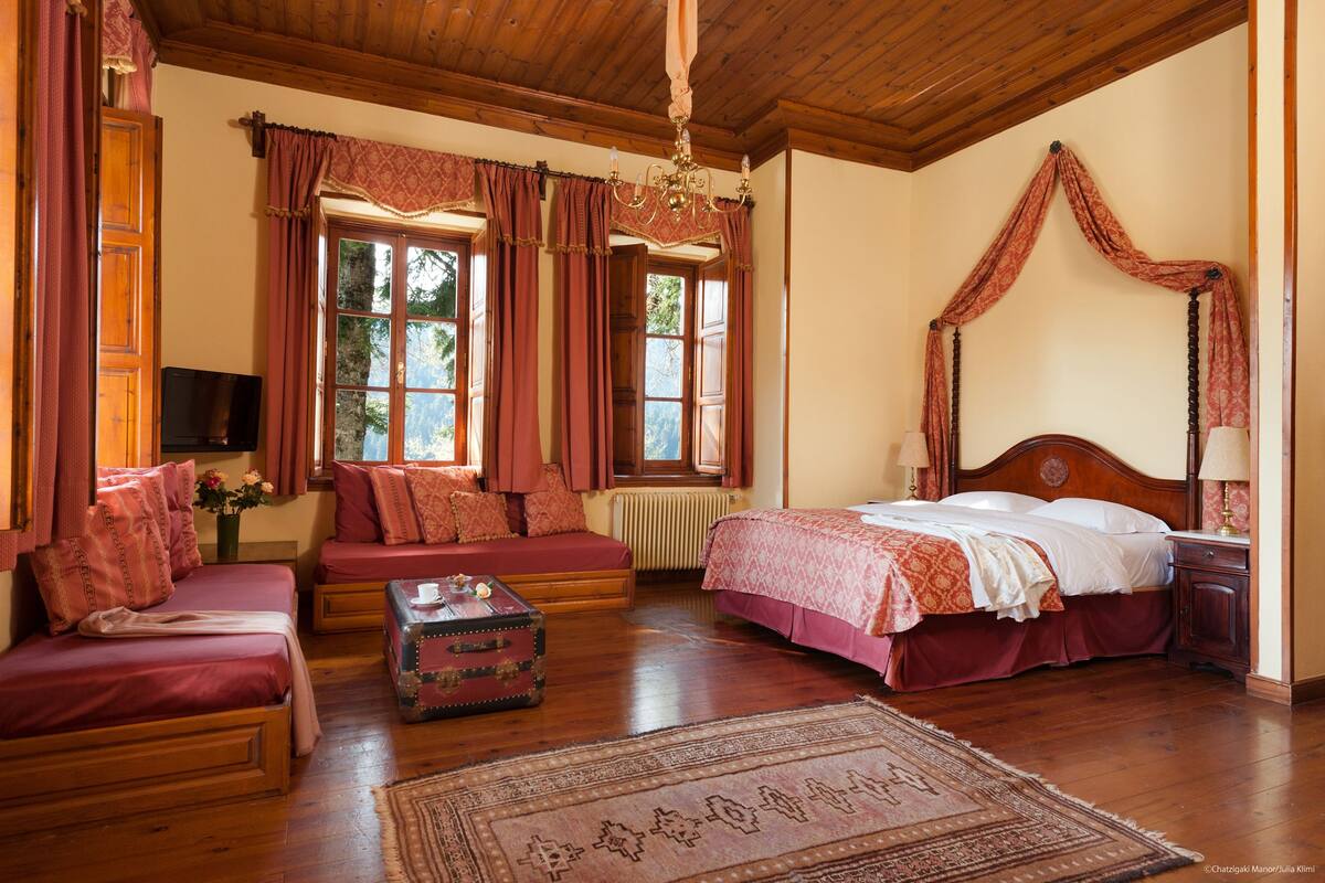 Ein Zimmer mit Holzbalkendecke, einem Himmelbett, einer Couch, einem kleinen Tisch und einem Koffer.