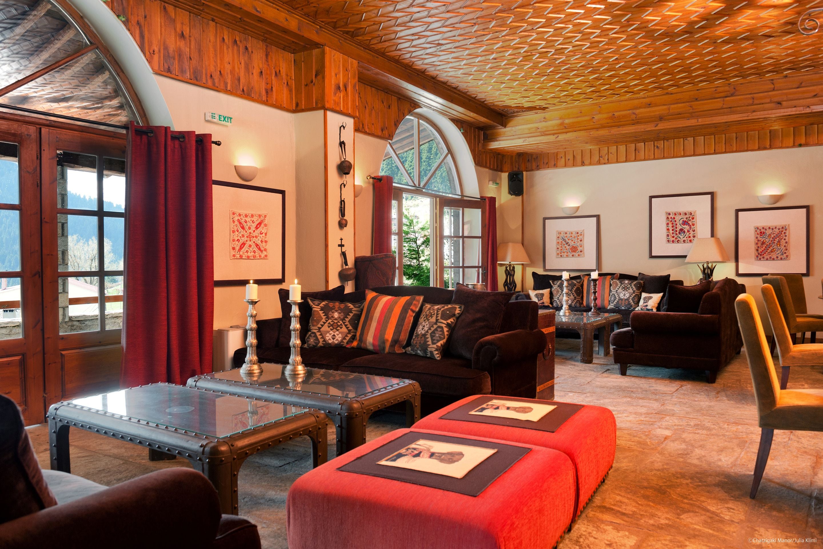 A cozy room with a wooden ceiling, a large window, and a fireplace. There are two sofas and a table with a candle.