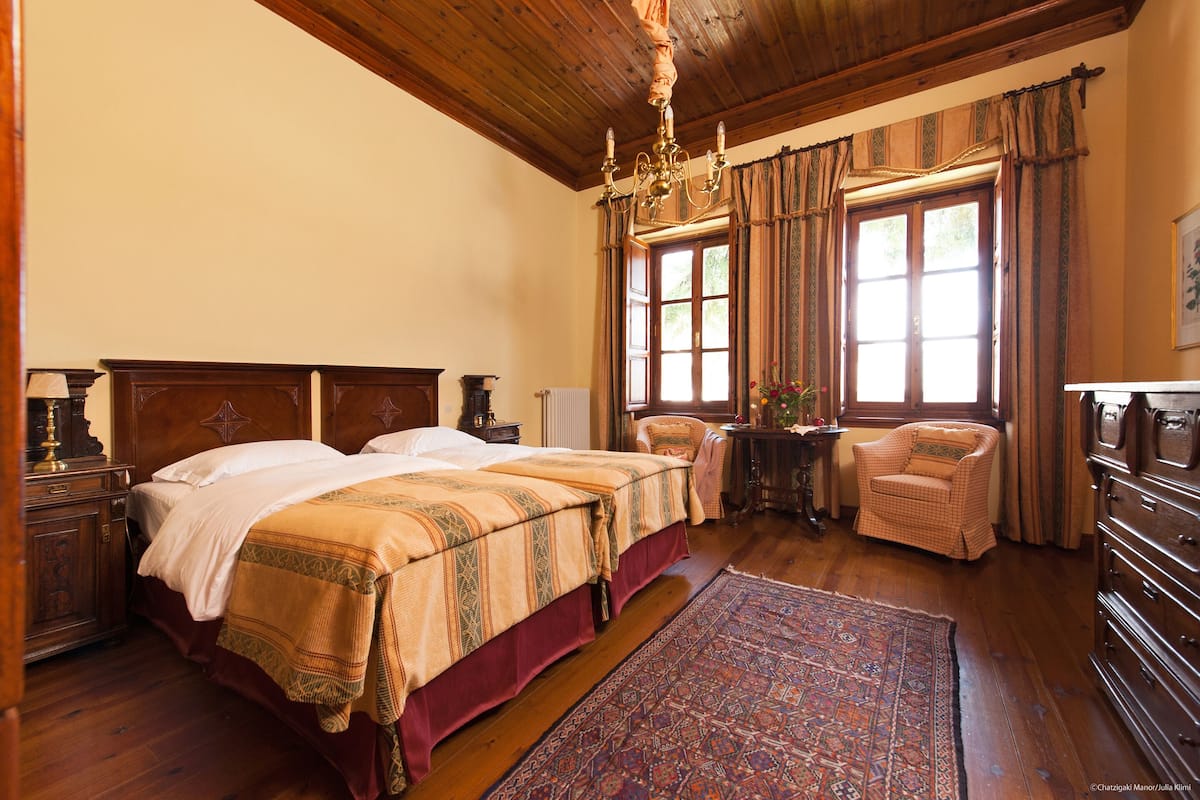 Ein traditionell eingerichtetes Schlafzimmer mit einem großen Bett, einem Sessel, einer Kommode und einem Fenster mit Vorhängen.