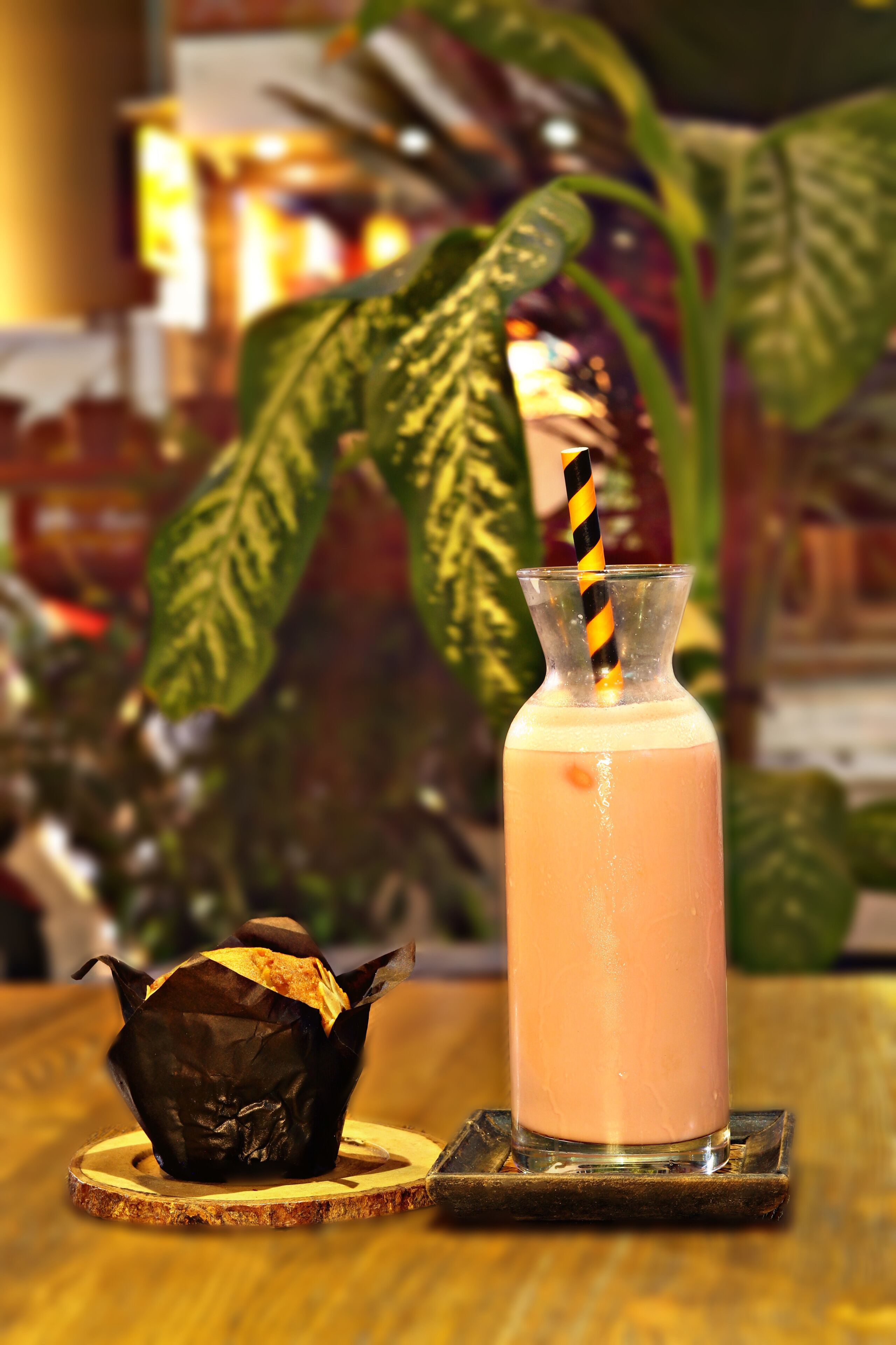 Un cóctel en un vaso con una pajita a rayas, un dulce cubierto de chocolate sobre un posavasos de madera, y una planta al fondo.