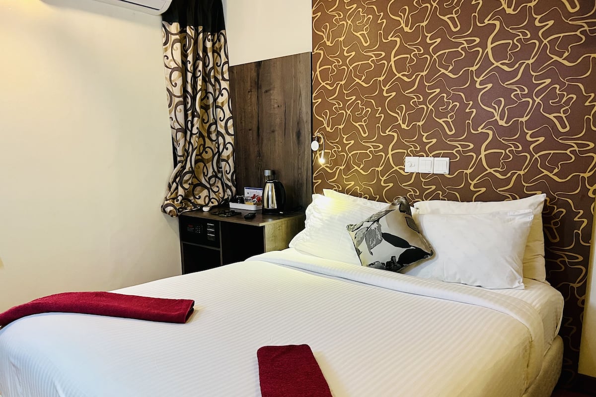 A hotel room with a neatly made bed, a bedside table with a coffee maker, and a patterned wall.