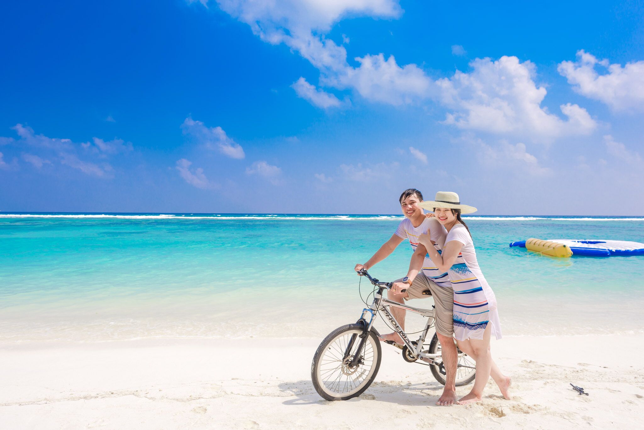 Una pareja en la playa con una bicicleta, agua azul cristalina y cielo despejado.