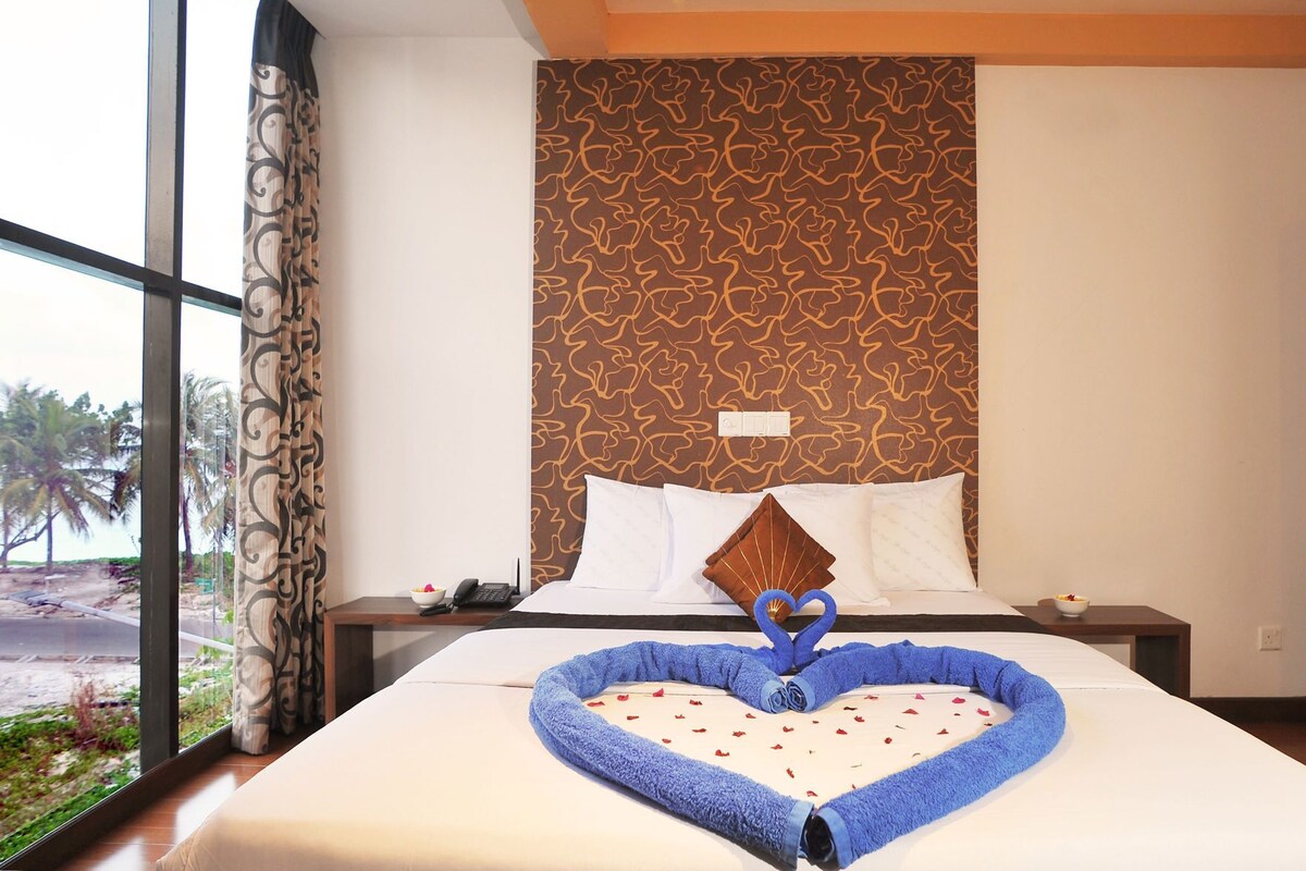 Una habitación de hotel con una cama grande, una toalla con forma de corazón sobre la cama y vistas a palmeras fuera.