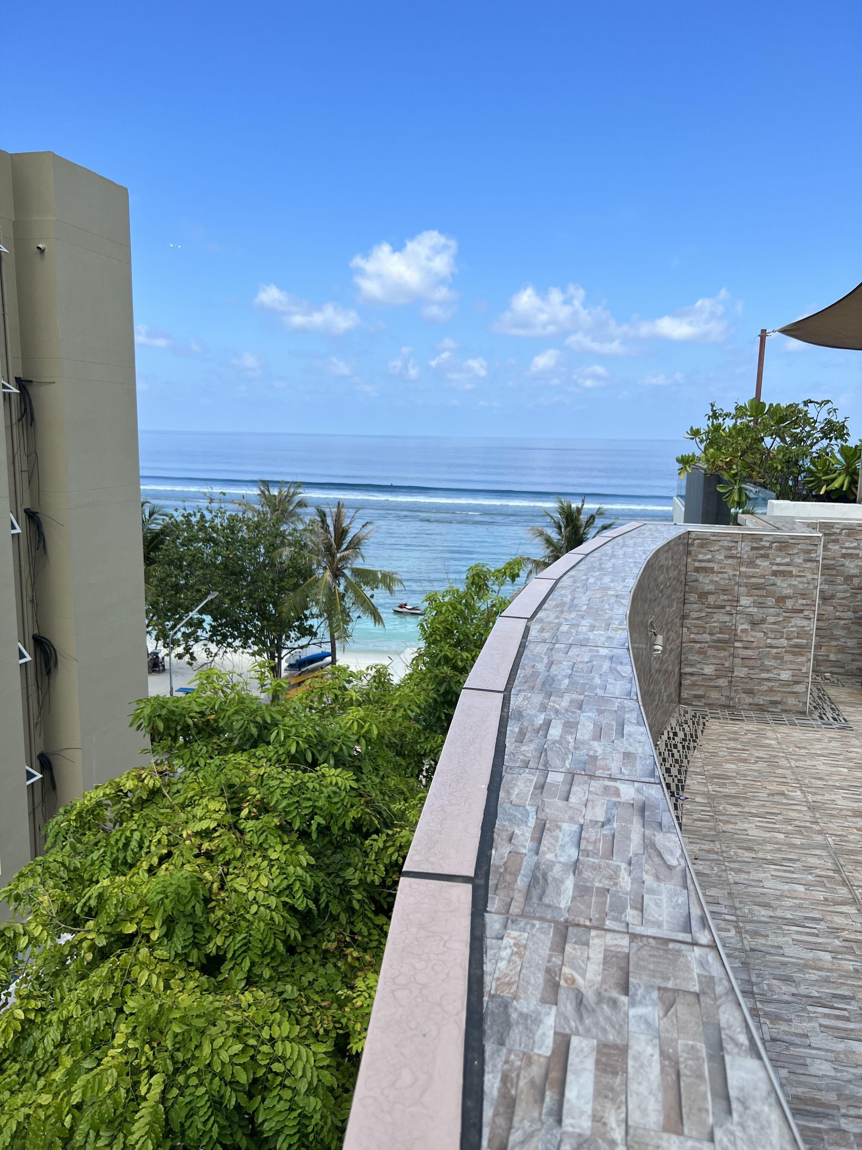A balcony with a view of the ocean, a stone wall, and some greenery.