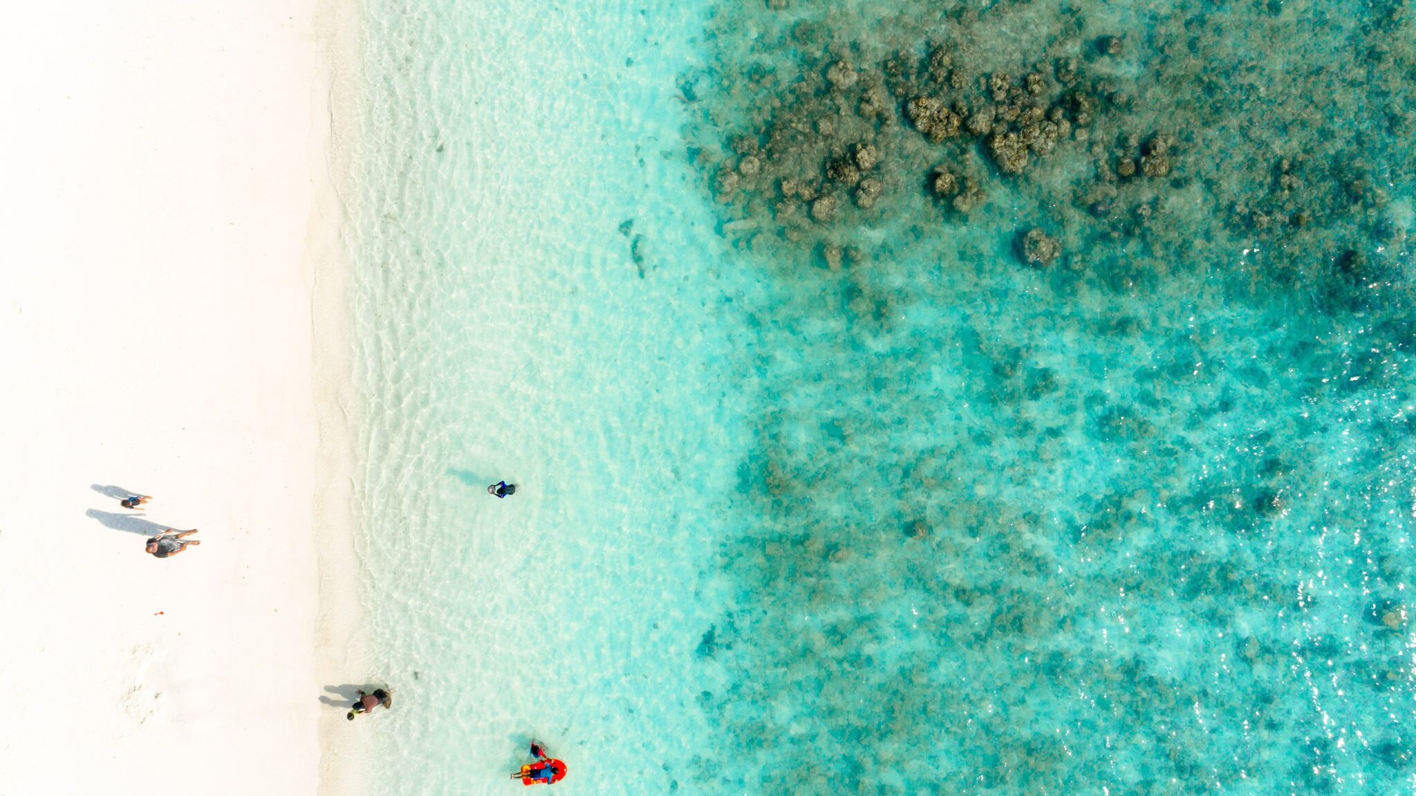 An aerial view of a beach with clear turquoise water, white sand, and a few people.