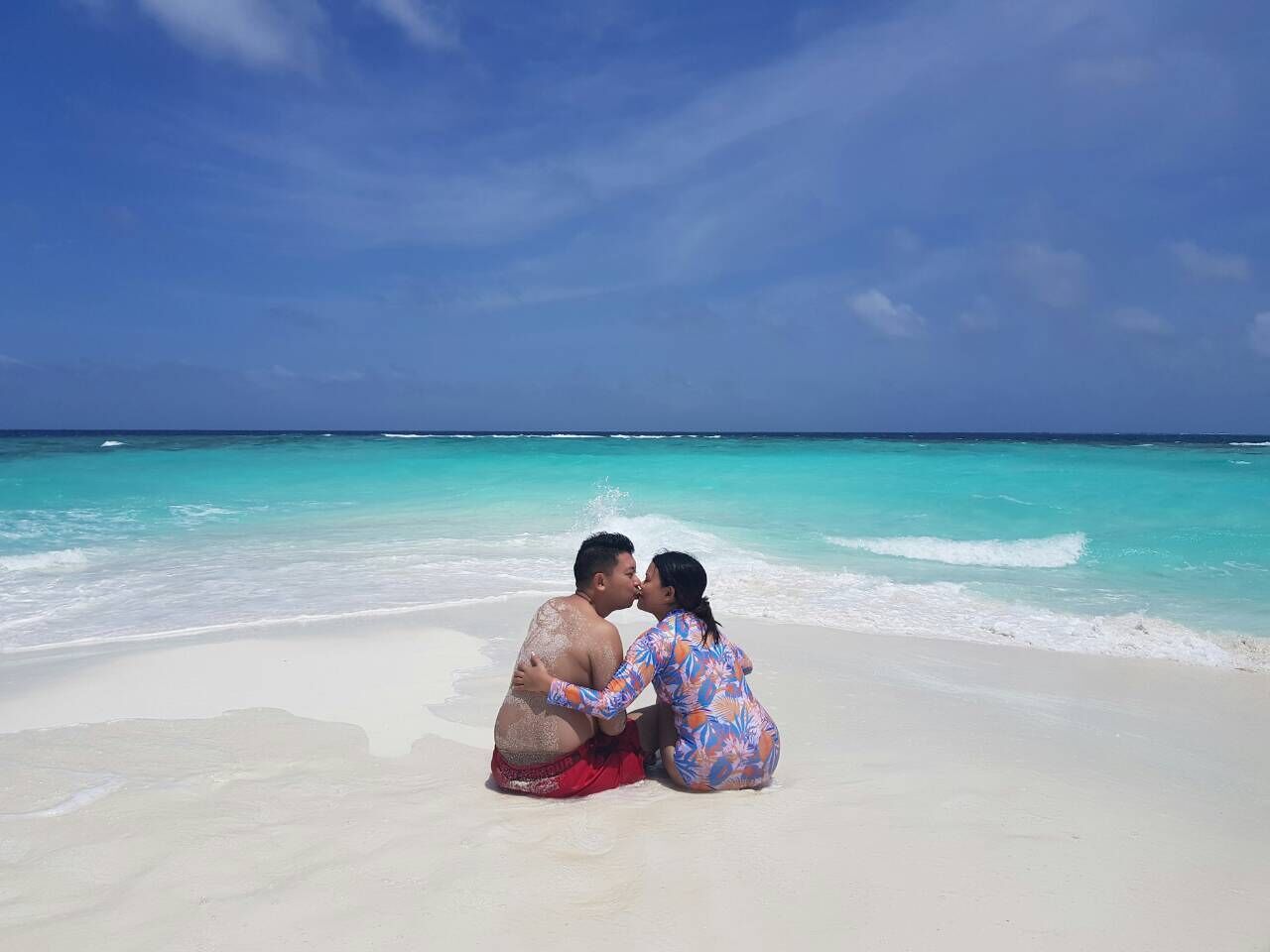 A couple sitting on a beach with clear turquoise water and a clear sky.