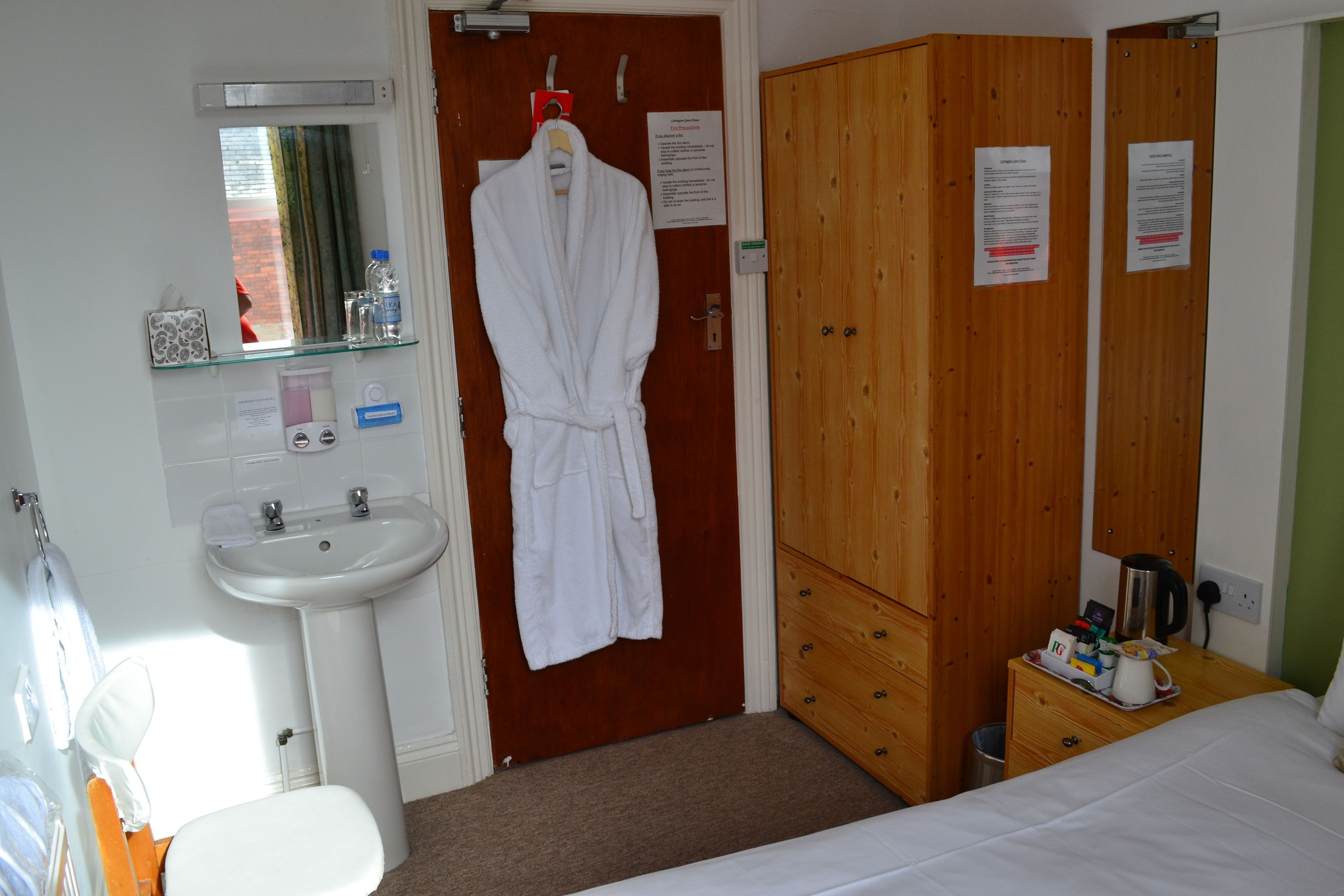 A hotel room with a white sink, a mirror, a wooden wardrobe, a bedside table with a kettle, and a bed.