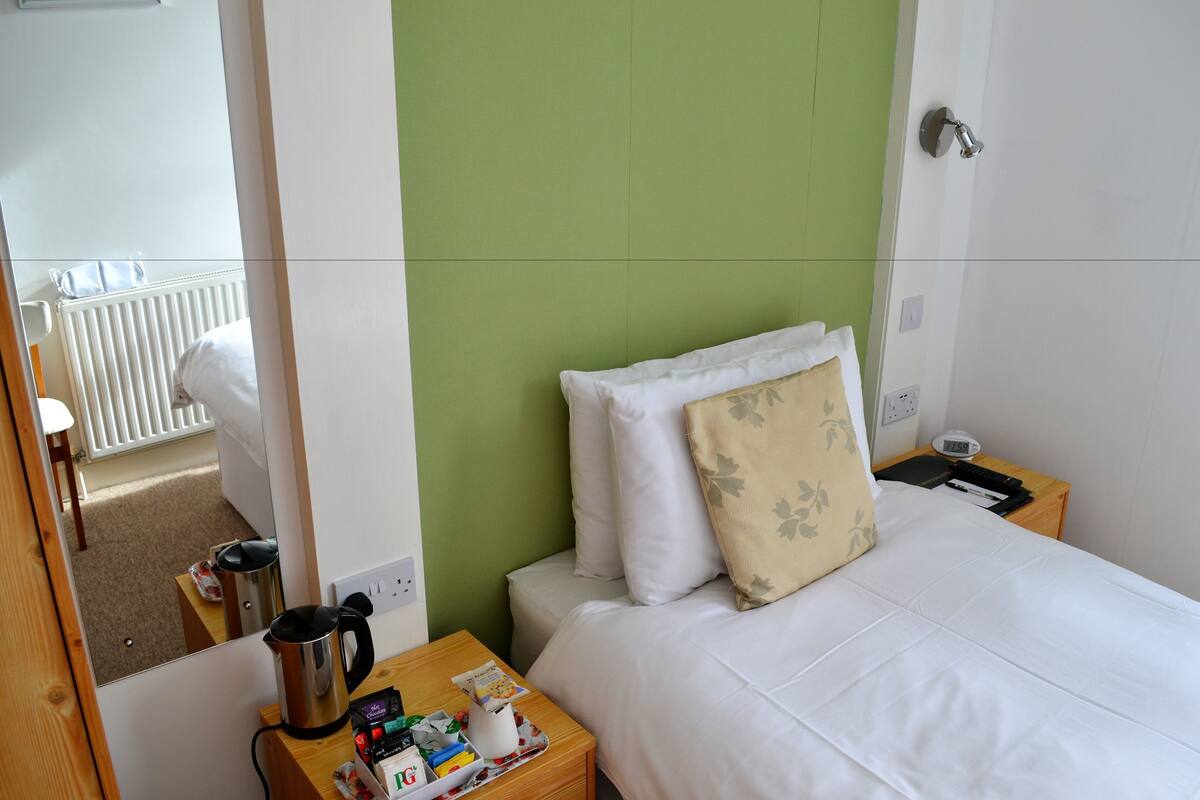 Ein Hotelzimmer mit Bett, Nachttisch mit Wasserkocher, kleinem Tisch mit Snacks sowie Blick auf einen weißen Heizkörper und einen Stuhl im Hintergrund.