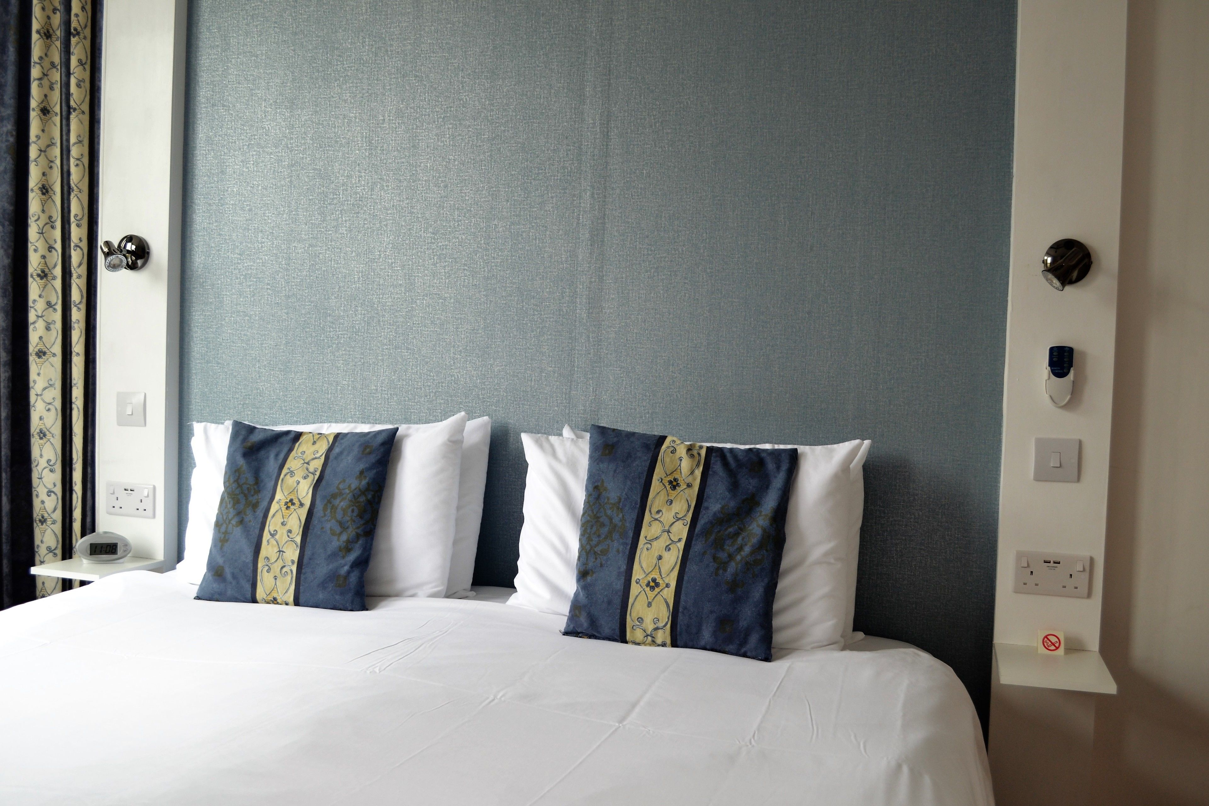 A hotel room with a bed, two decorative pillows, a nightstand with a clock, and a wall-mounted light switch.