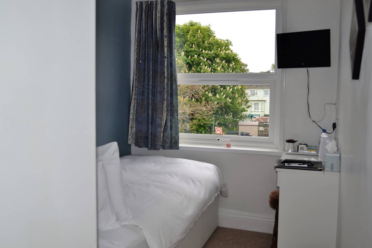 Ein Schlafzimmer mit einem Einzelbett, einem Fenster mit Vorhängen, einem an der Wand montierten Fernseher und einem kleinen Tisch mit Gegenständen darauf.