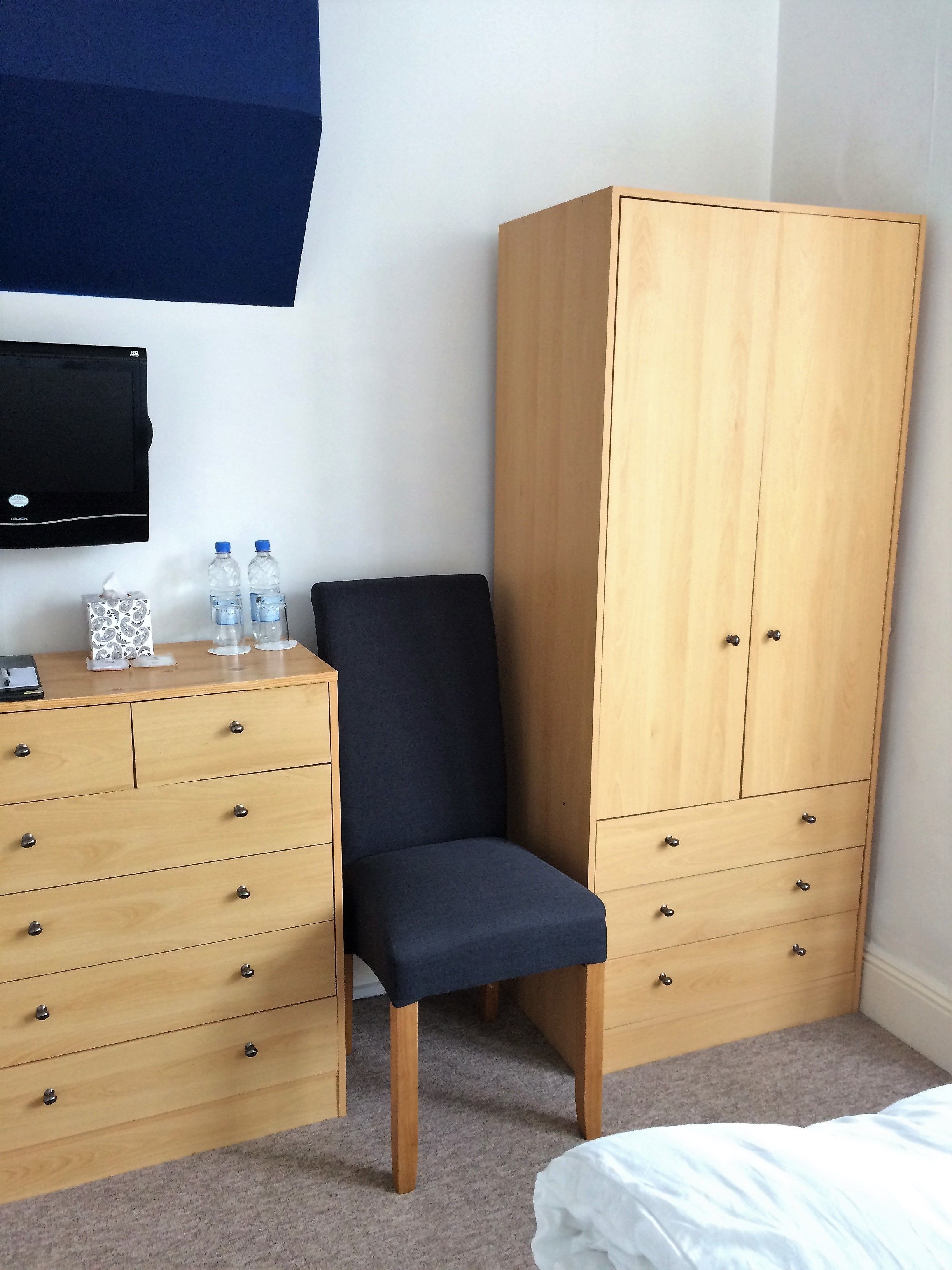 Ein Schlafzimmer mit Holzschrank, Stuhl, Bett, Fernseher und kleinem Tisch mit Toiletteartikeln.