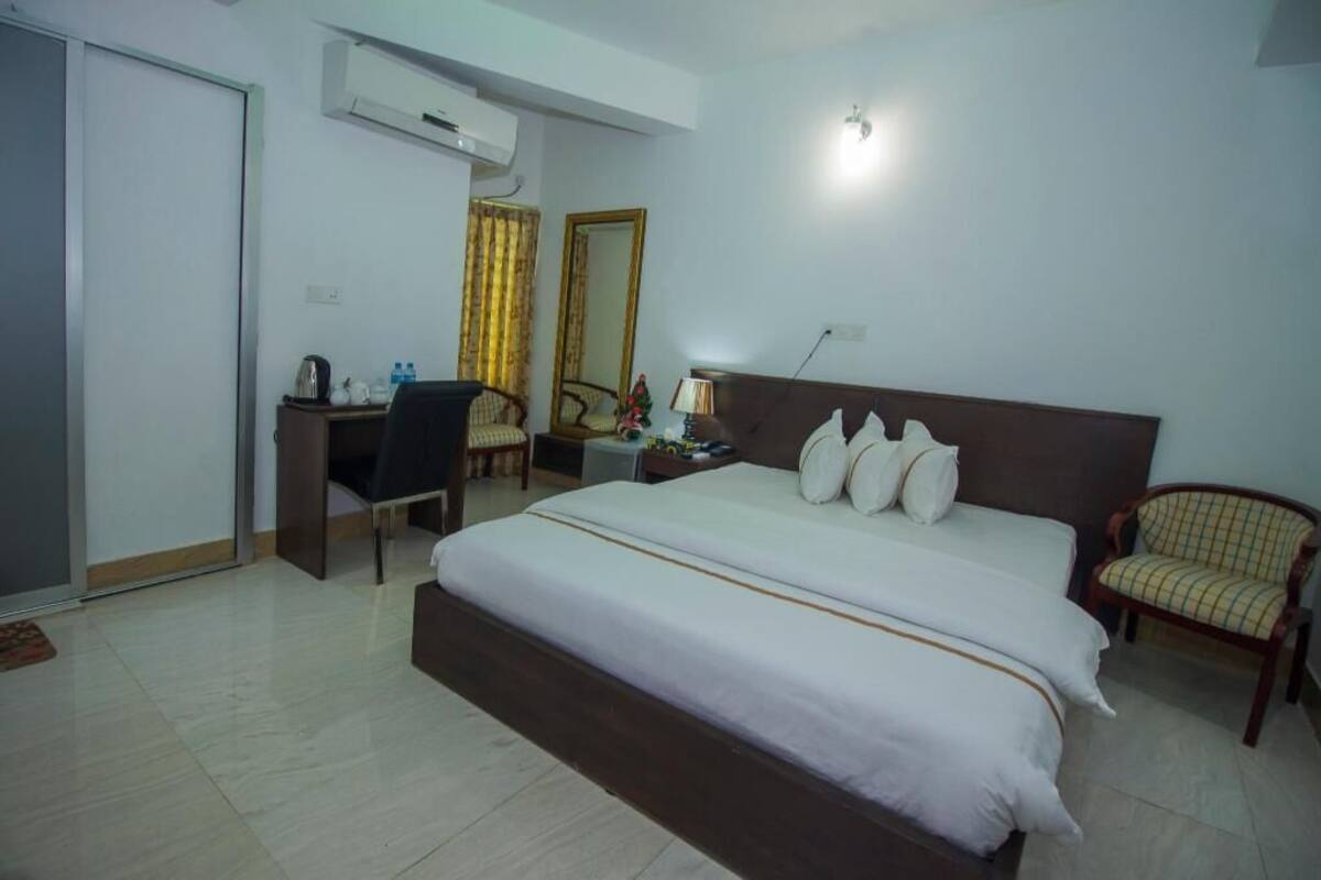 A hotel room with a bed, a desk, a chair, a mirror, and an air conditioning unit.