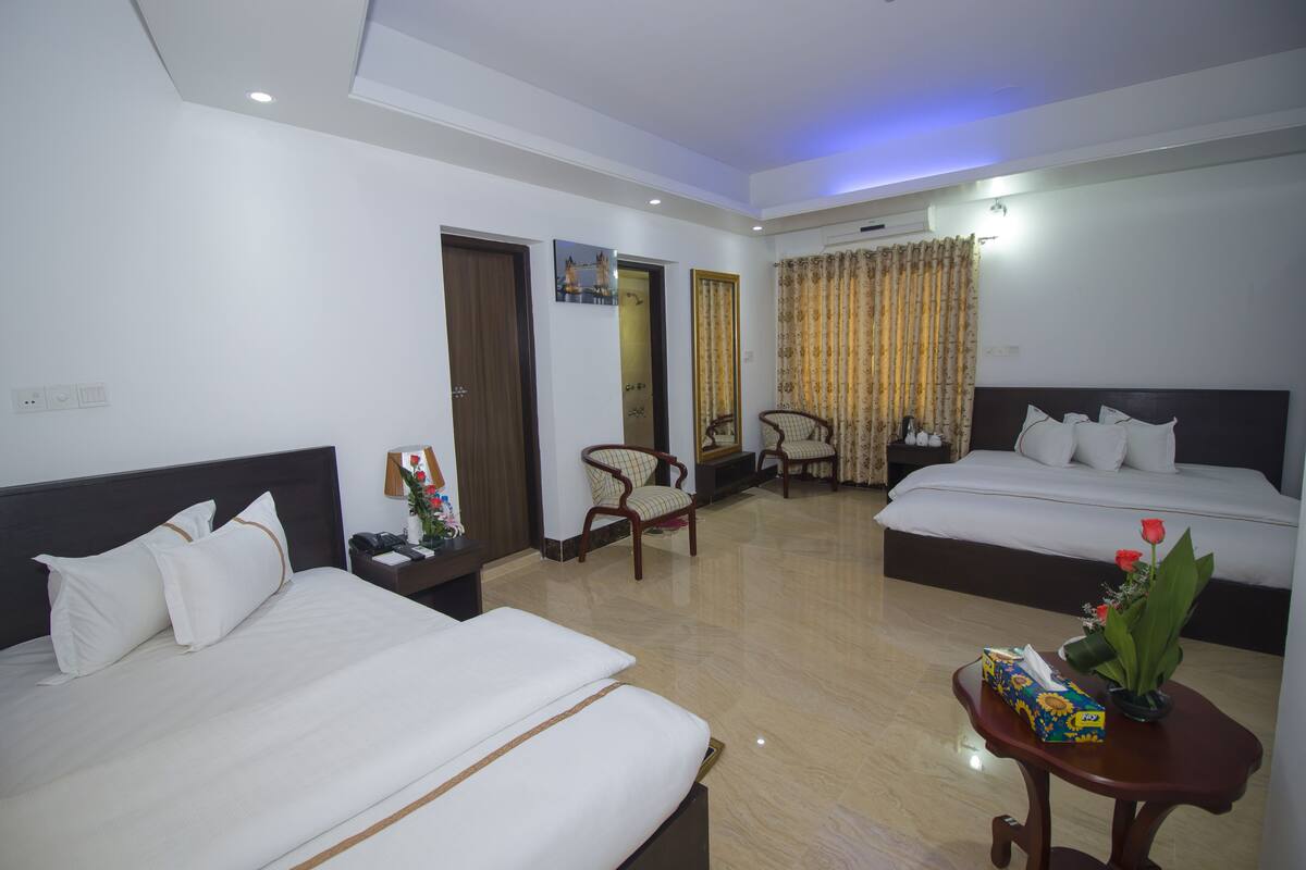 A hotel room with two beds, a wooden nightstand, a small table with a vase and a box, and a chair.