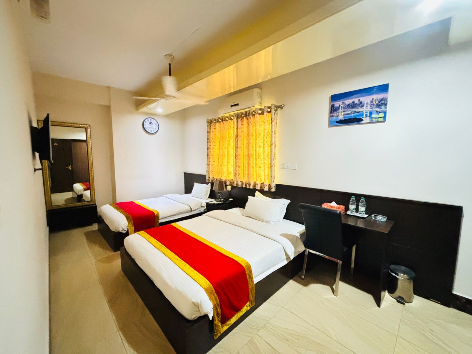 A hotel room with two beds, a desk, a chair, a mirror, a clock, and a picture on the wall.