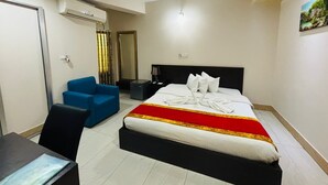 Quarto de hotel com cama, escrivaninha, cadeira, ventoinha no teto, aparelho de ar condicionado, espelho e um quadro na parede.
